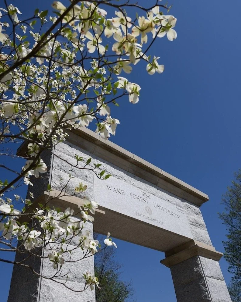 A stone archway with inscriptions for Wake Forest University and the dates 1834 and 2023, framed by blossoming white flowers on a bright, clear day.