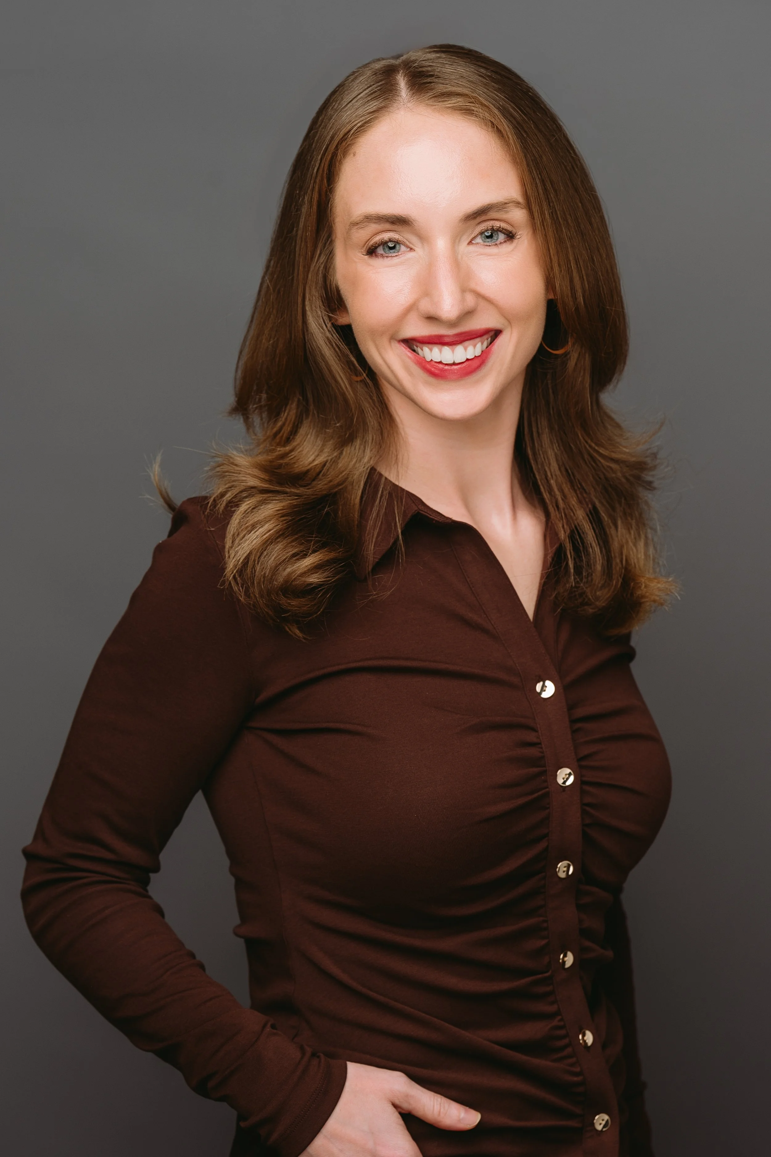 A woman with light skin, brown hair, and blue eyes smiling, dressed in a dark brown, long-sleeve blouse with buttons, standing against a gray background.