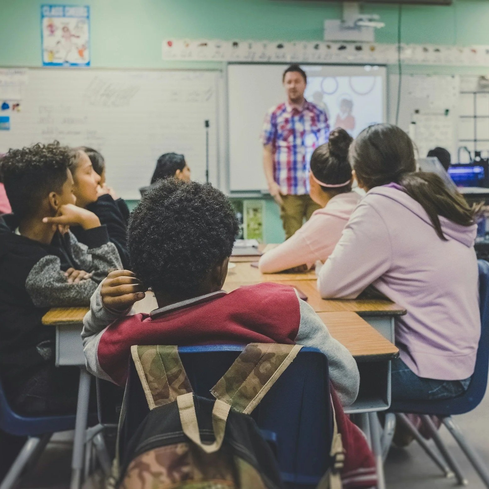 Classroom with students sitting at desks listening to a teacher at the front, who is standing near a whiteboard and a projector screen with a digital presentation.