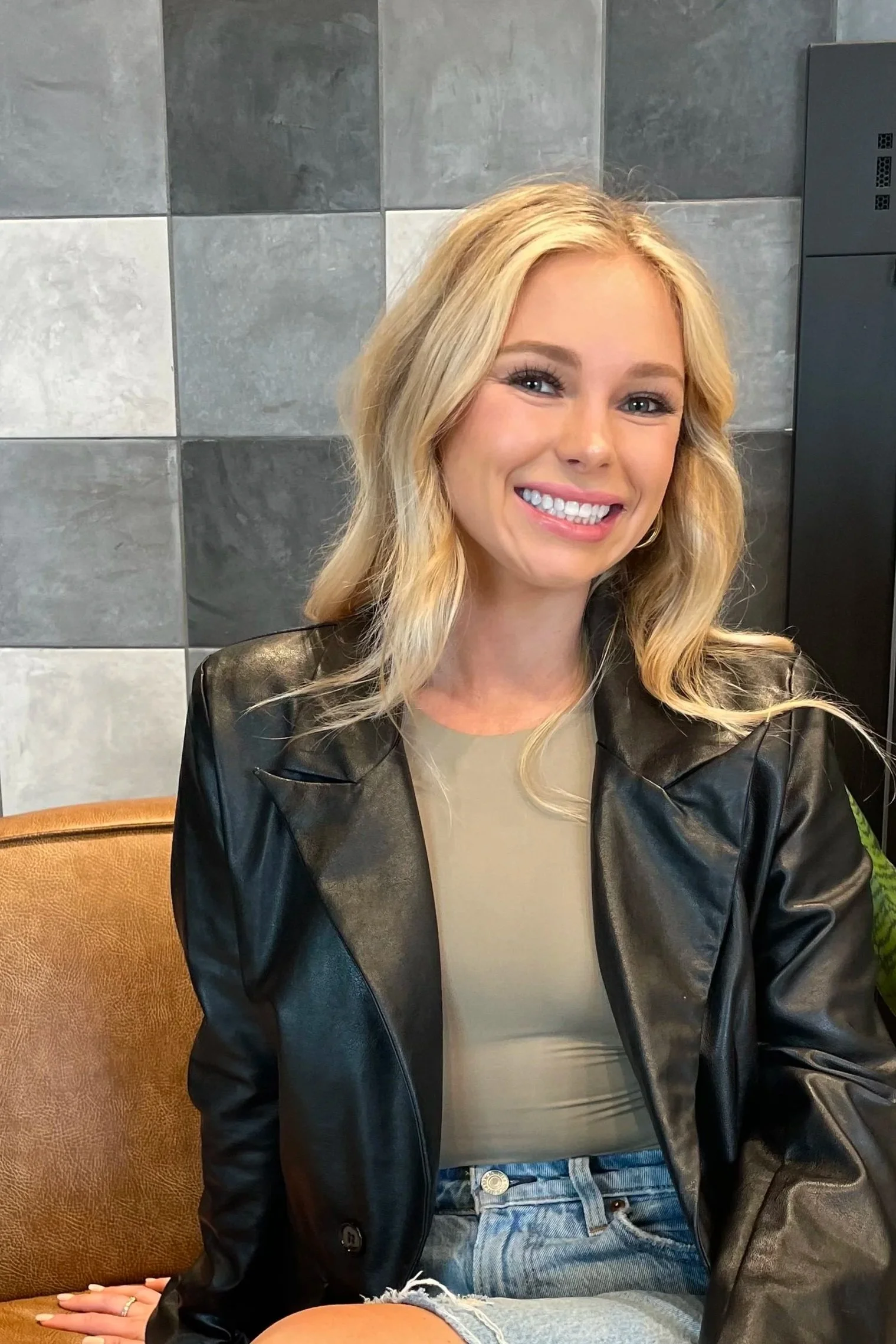 A young woman with blonde wavy hair smiling, wearing a black leather jacket over a beige top and denim shorts, seated against a tiled wall.