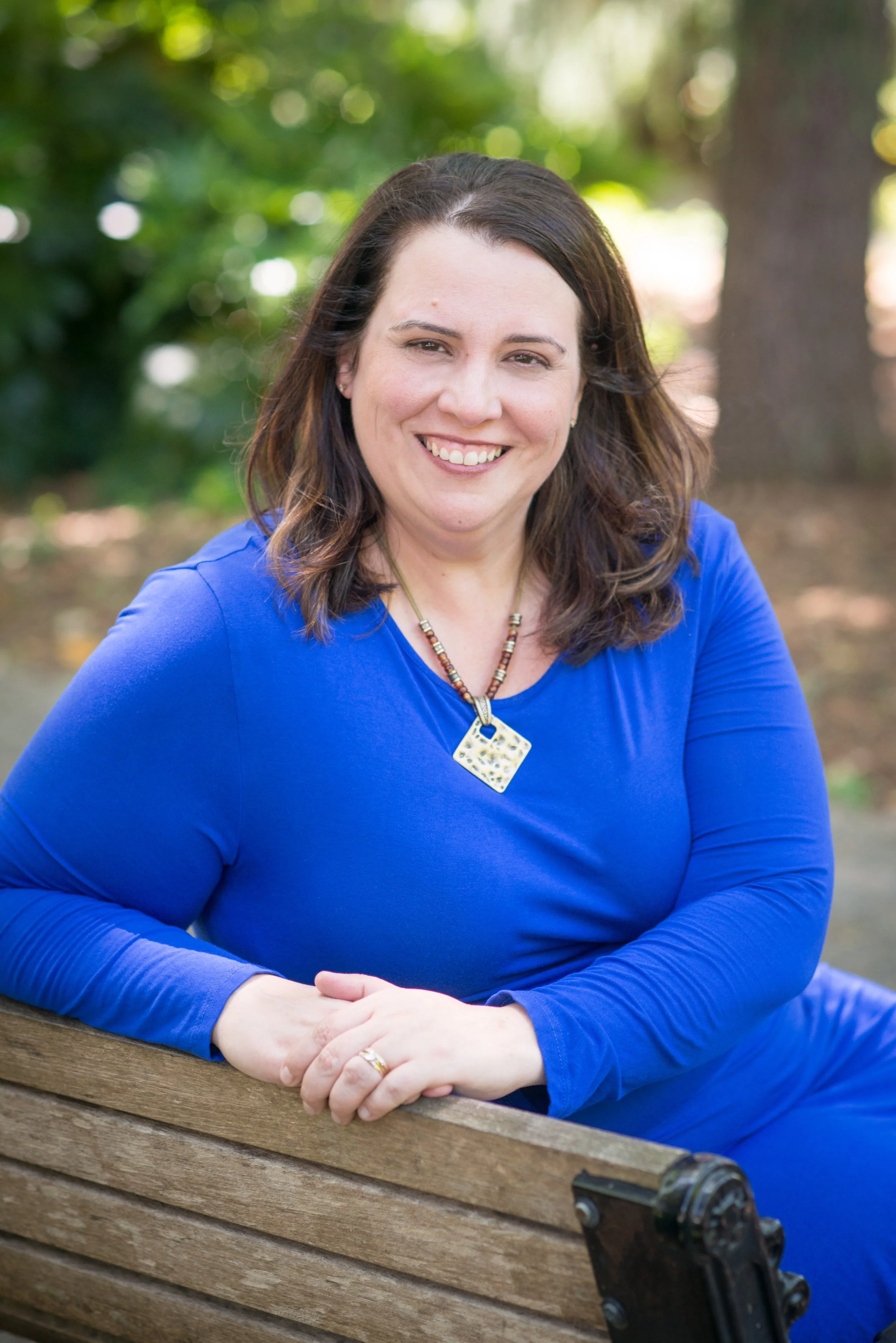 A woman with shoulder-length brown hair, smiling, sitting on a wooden park bench wearing a bright blue dress and a beaded necklace, outdoors with greenery in the background.