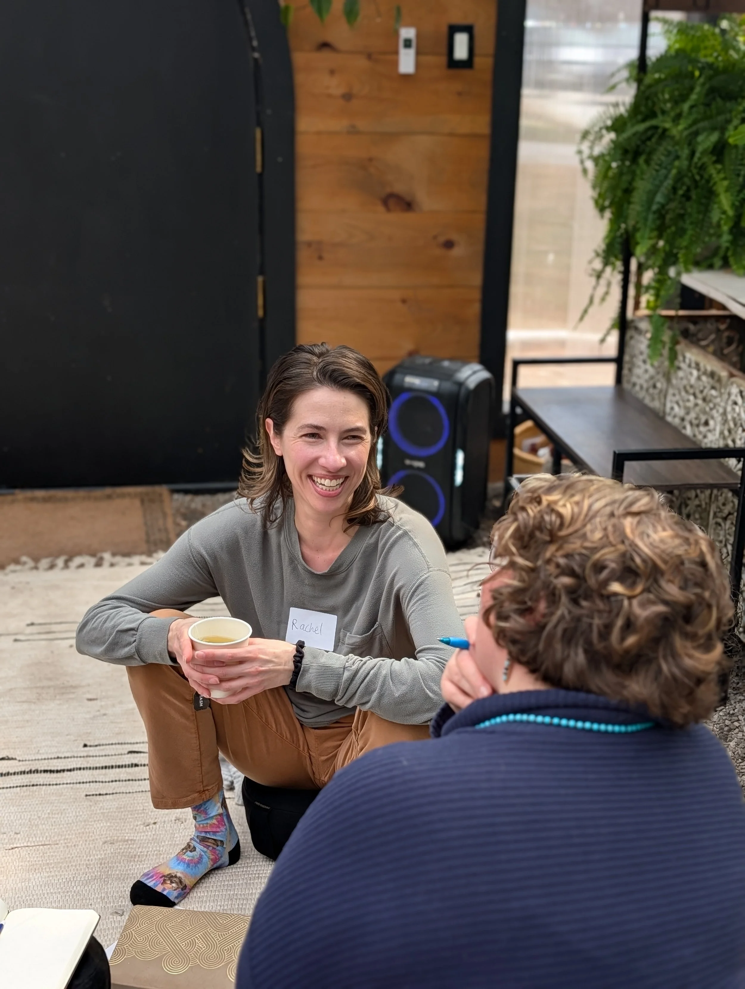 Two women sitting on the floor having a conversation, one woman is smiling and holding a cup, the other woman is holding a pen. They are in a cozy indoor space with wooden walls, plants, and a speaker in the background.