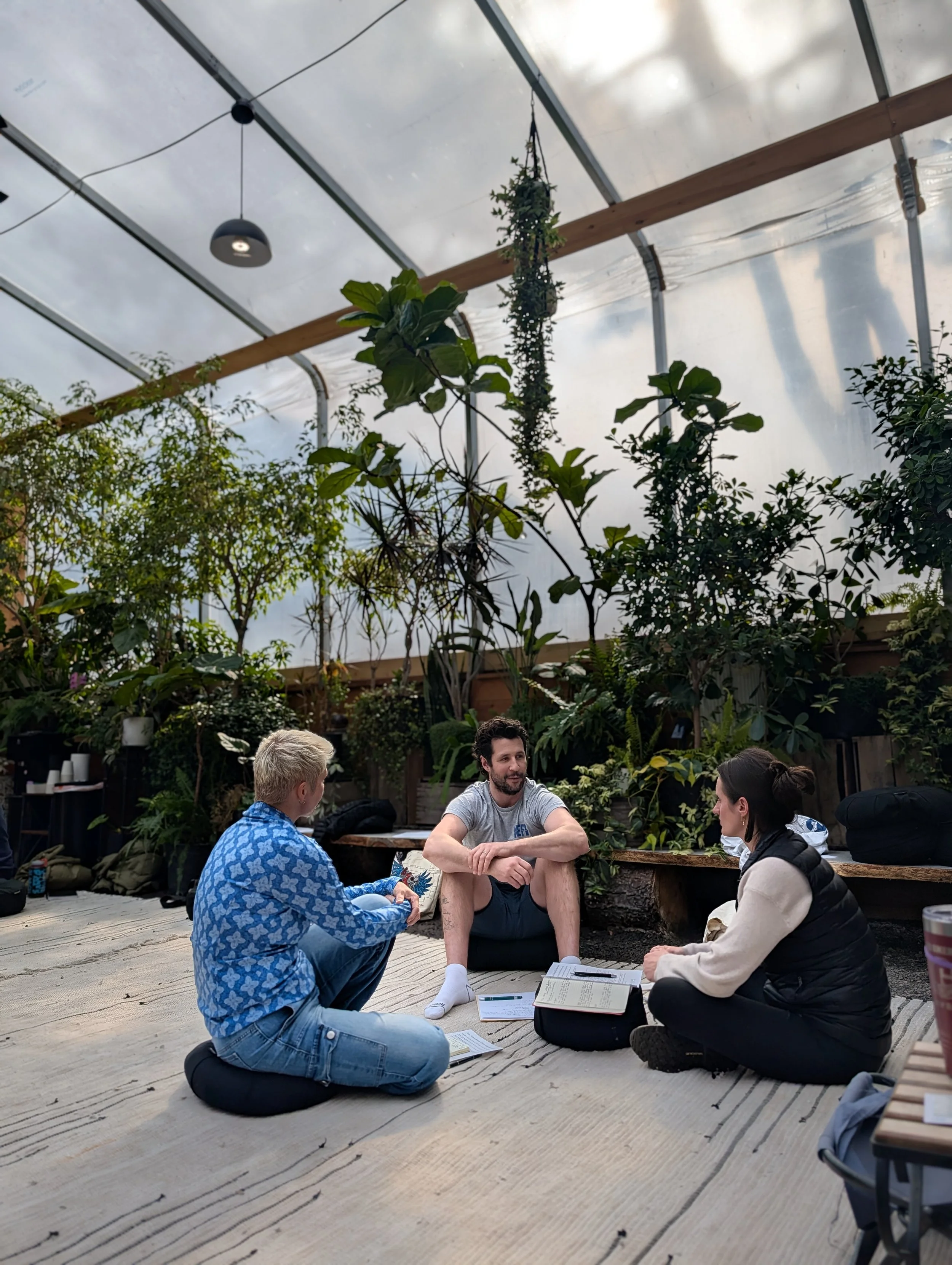 Three people sitting on the floor of a greenhouse, engaged in conversation, with notebooks and papers around them, surrounded by lush green plants and large windows overhead.