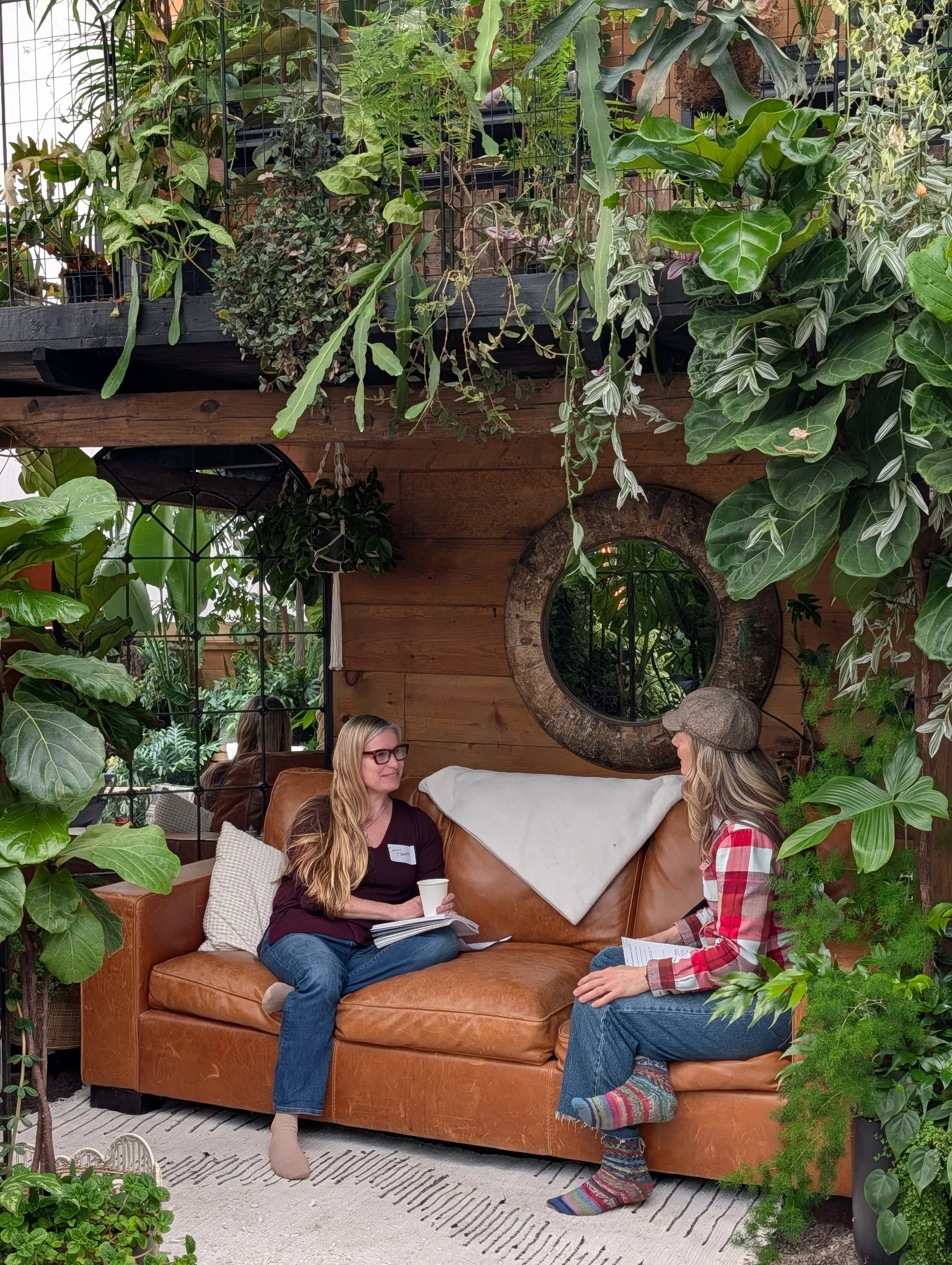 Two women sitting on a leather couch in a cozy, plant-filled indoor space, engaged in conversation. The woman on the left has long blond hair, glasses, and wears a dark top with jeans, holding a cup. The woman on the right wears a plaid shirt, a hat, and colorful socks, holding papers. The room features a wooden wall, a round mirror, and various lush green plants surrounding them.