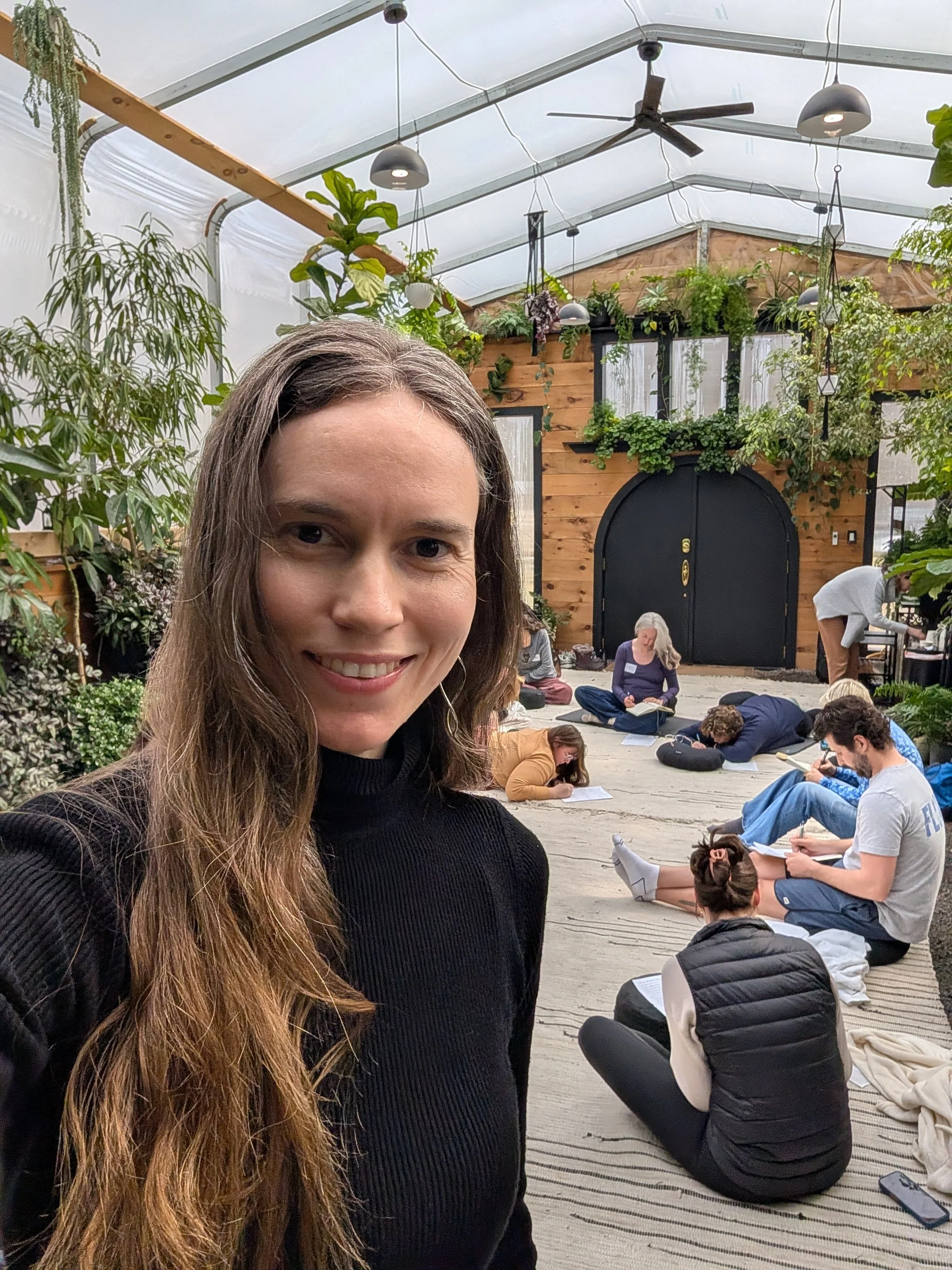 A woman with long brown hair smiling at the camera inside a greenhouse with people sitting on the floor, writing on papers, surrounded by green plants and wooden walls.