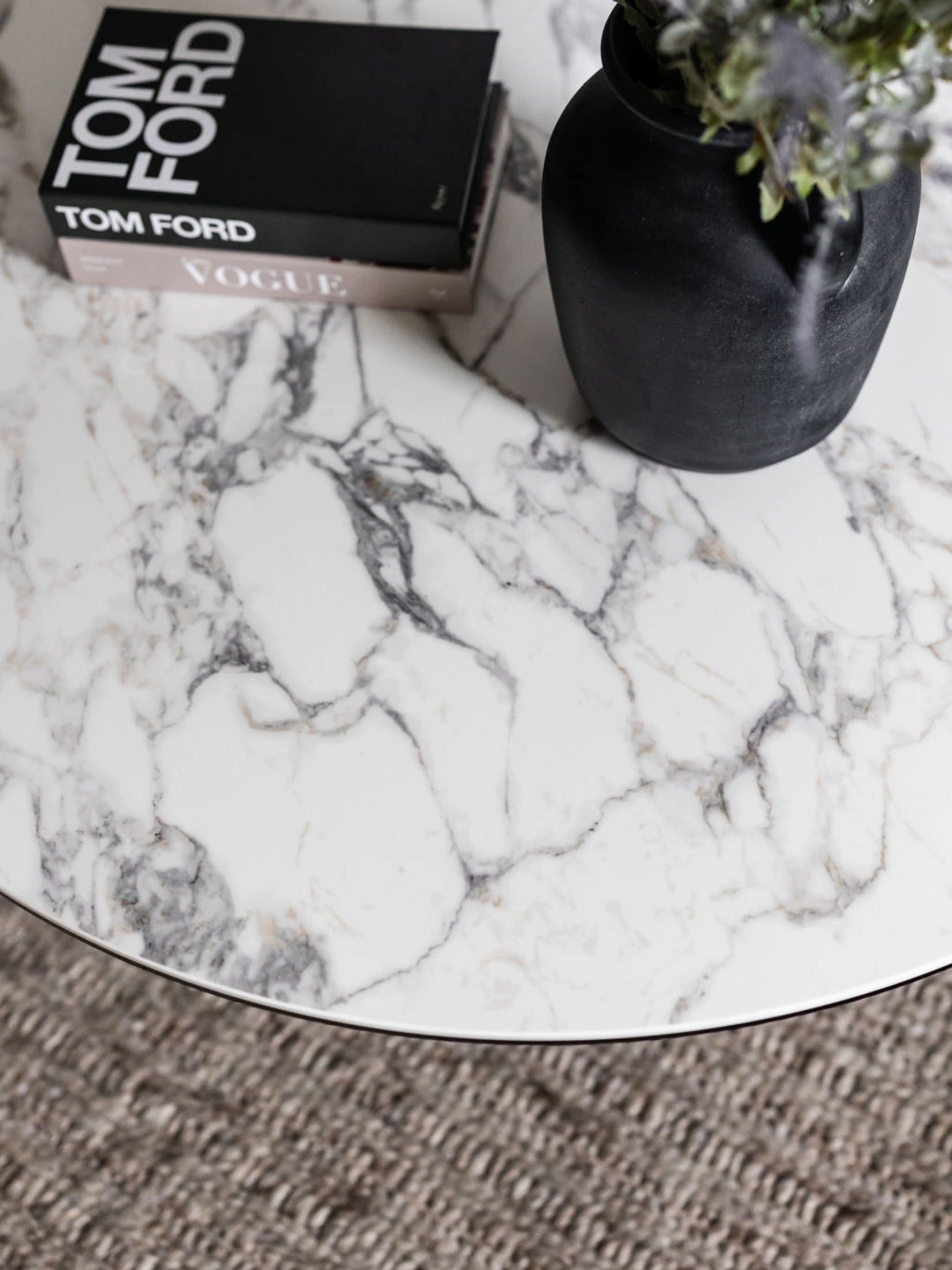 Close-up photo of a white marble table with gray veins, featuring two stacked books titled "Tom Ford" and "Vogue" and a black vase with some green plant leaves.