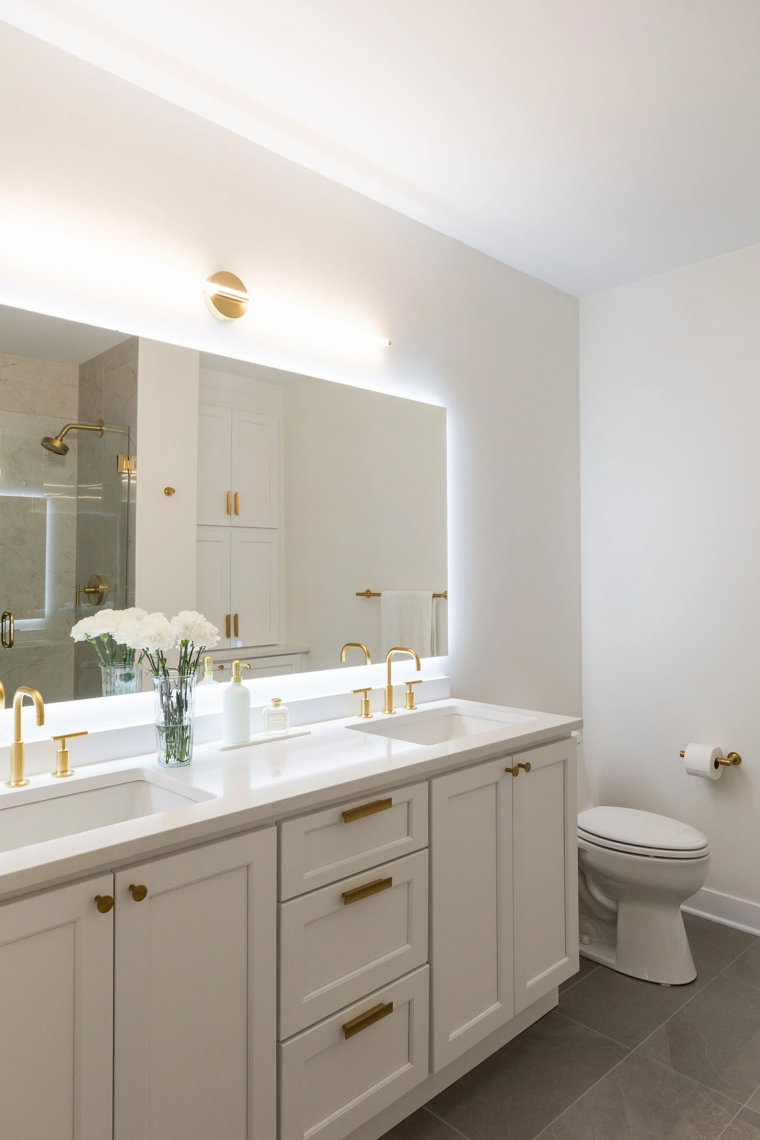 A clean, modern bathroom with white cabinetry, a double sink with gold fixtures, a large mirror, white walls, a white toilet, and a shower with a gold showerhead. Decor includes a vase of white flowers and toiletries on the counter.