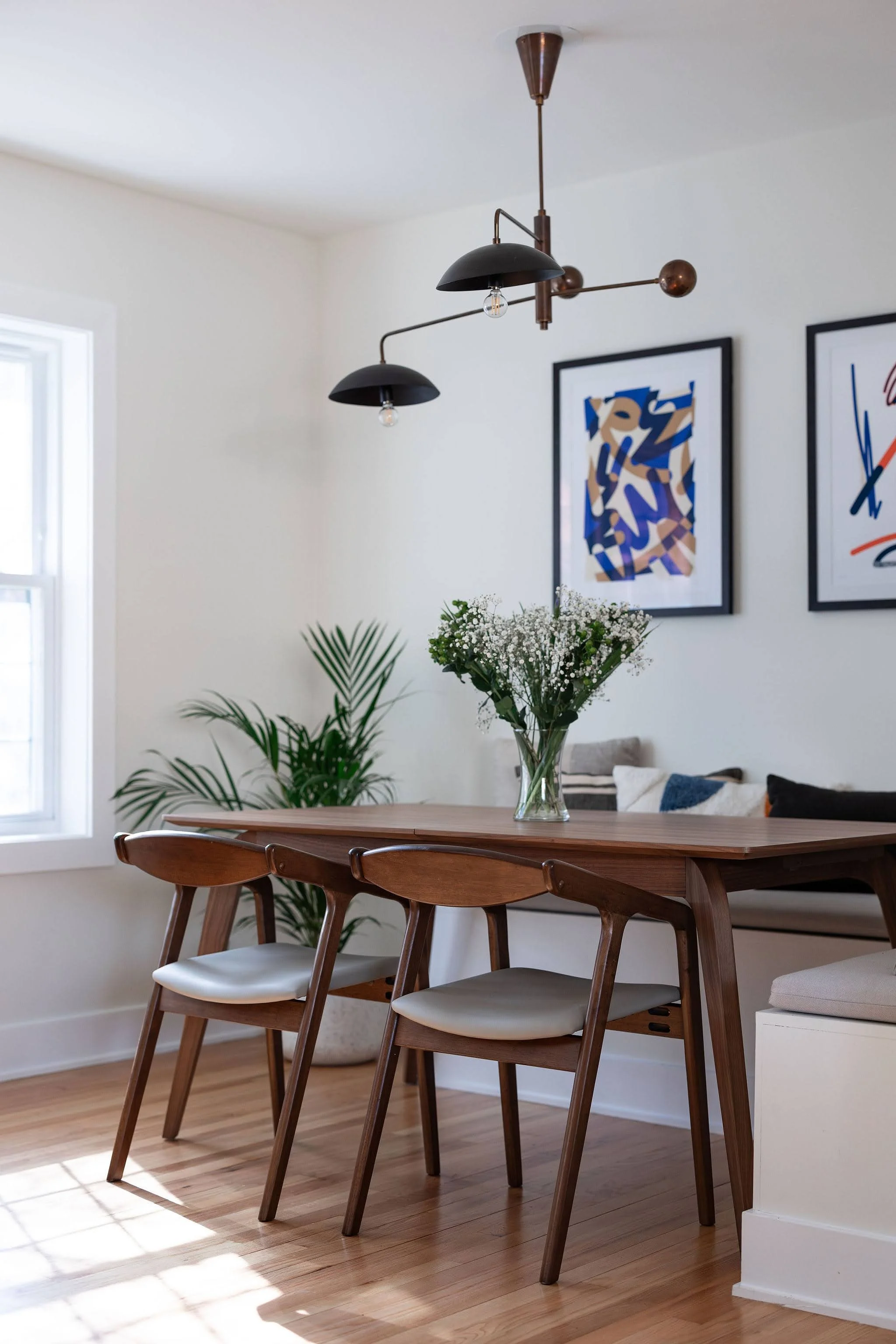 Dining area with wooden table, modern chairs, a glass vase with white flowers, black wall art, a window, and a black and brass ceiling light fixture.