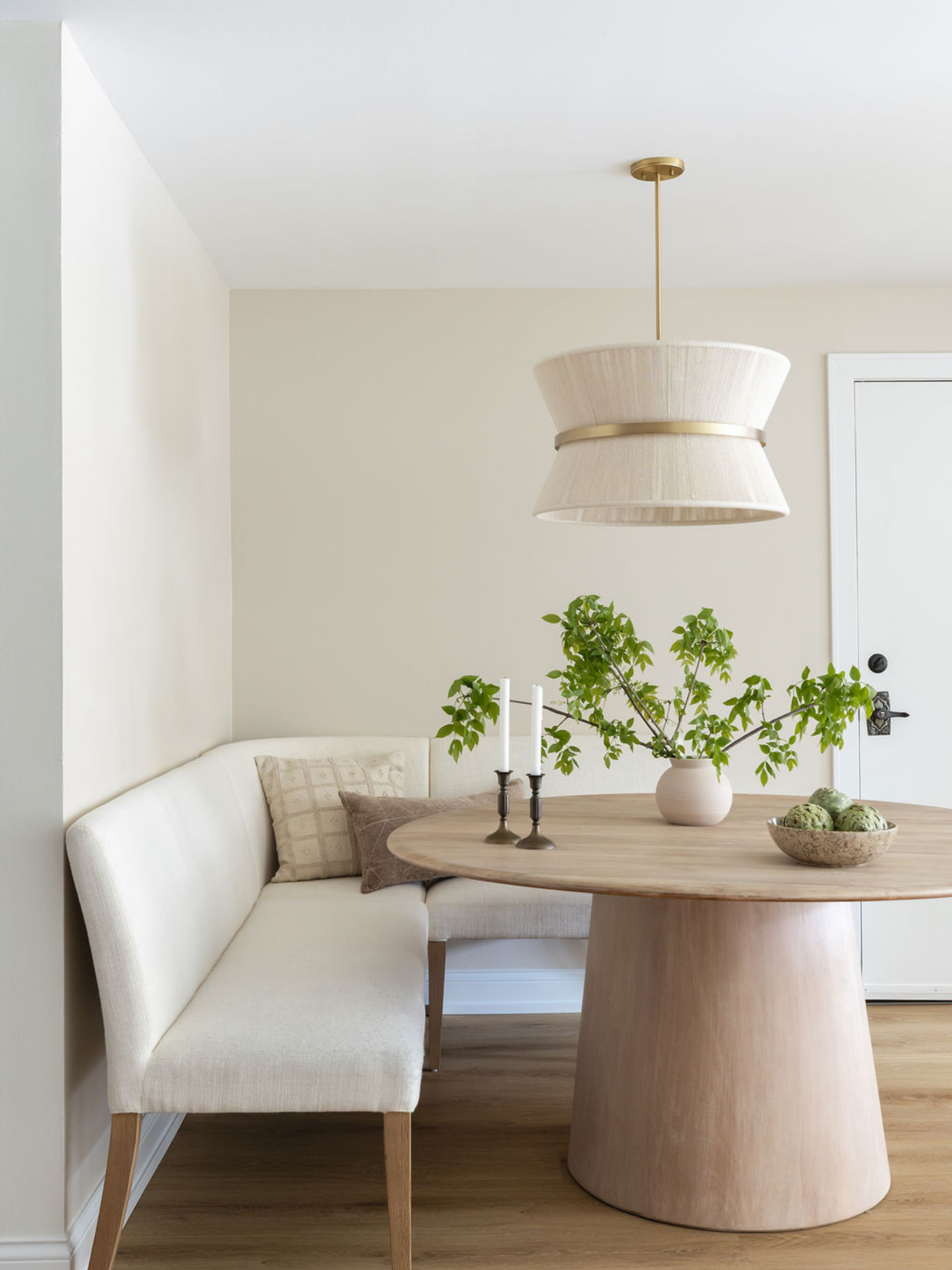 A cozy dining nook with a beige curved sofa, a round wooden table, a potted plant, a candelabra, a bowl of artichokes, and a white drum pendant light hanging from the ceiling.
