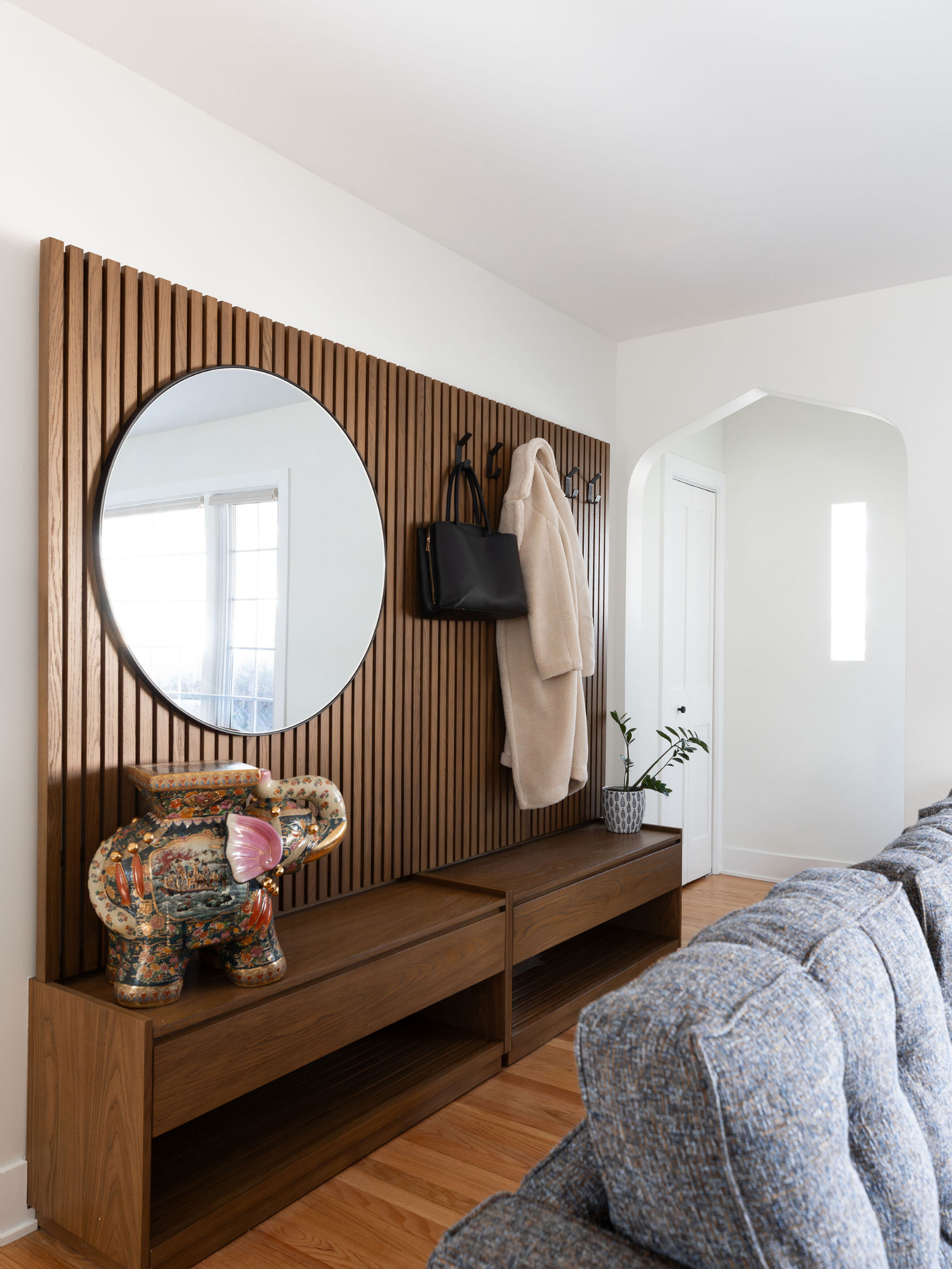 A wooden hallway accent wall with a round mirror, hooks holding a black bag and a beige coat, a potted plant on a wooden bench, a colorful ceramic elephant sculpture, and part of a blue and gray upholstered sofa in a living room.