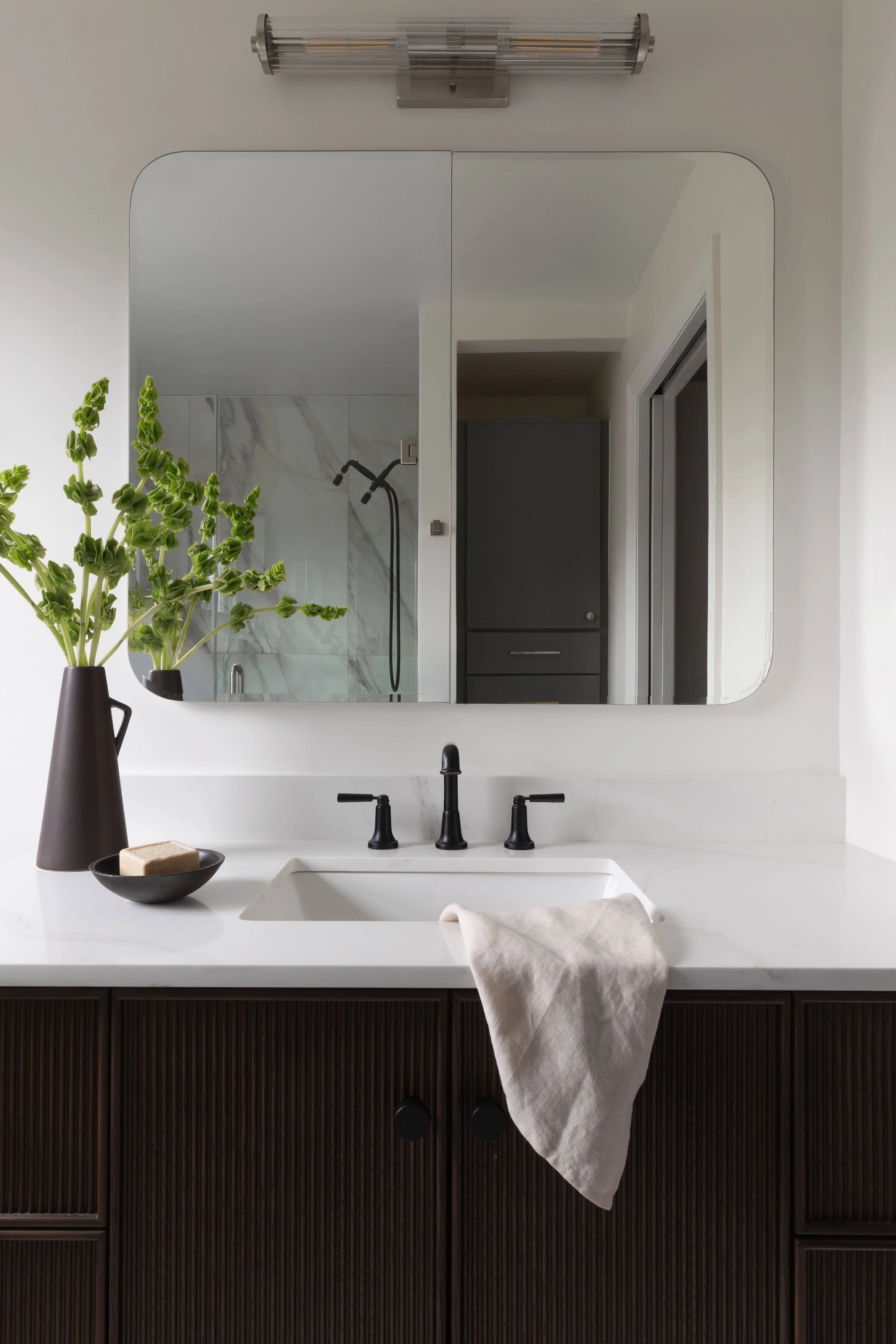Bathroom mirror showing a white sink with black faucet and dark wood vanity, a towel hanging on the front, a soap dish with soap, and decor like a vase with greenery. Outside the mirror, a shower with marble tiles and a black showerhead are visible.