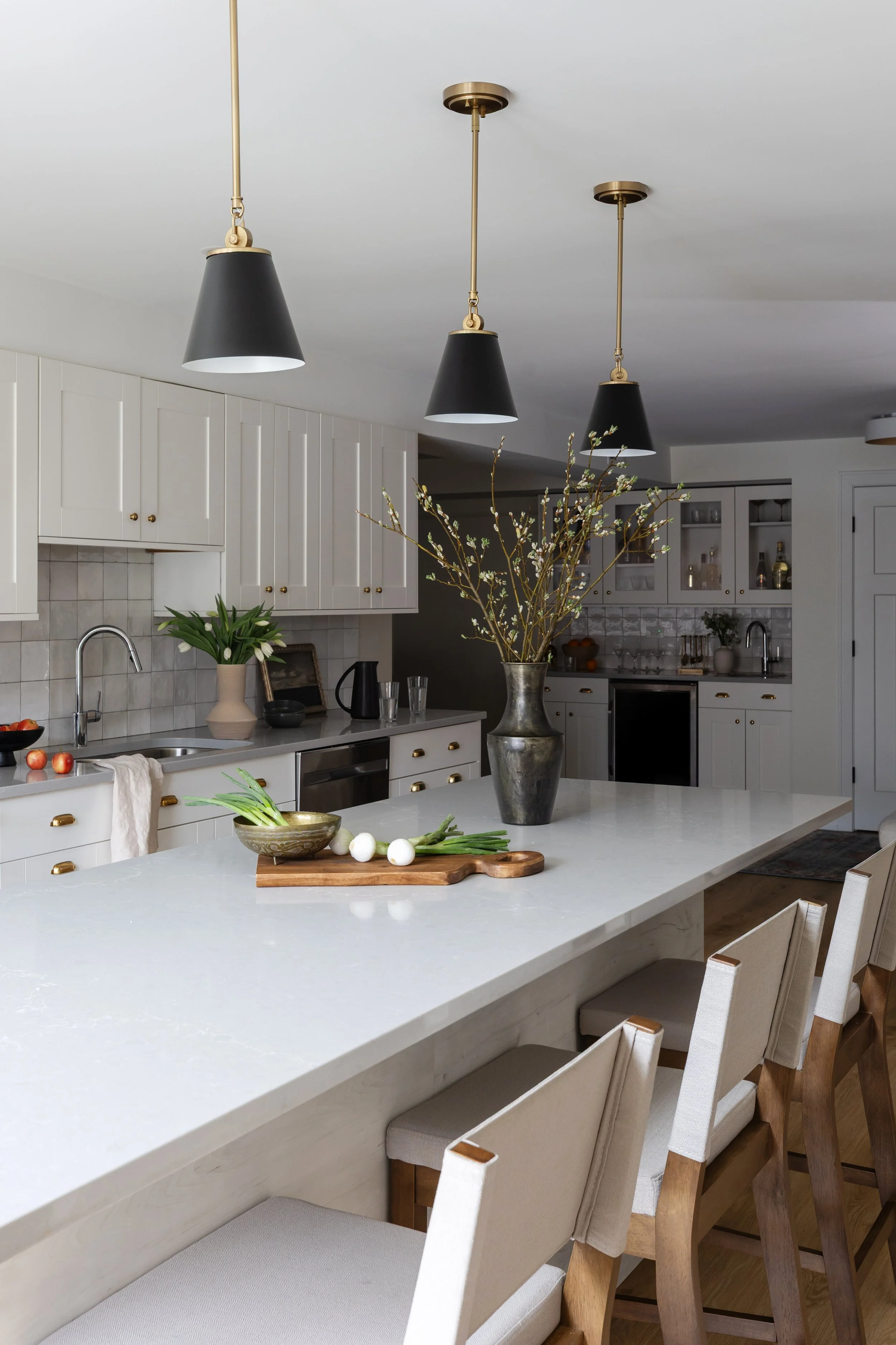 Modern kitchen with white cabinets, black pendant lights, a vase with branches on the island, a potted plant, and various kitchen utensils and decor.