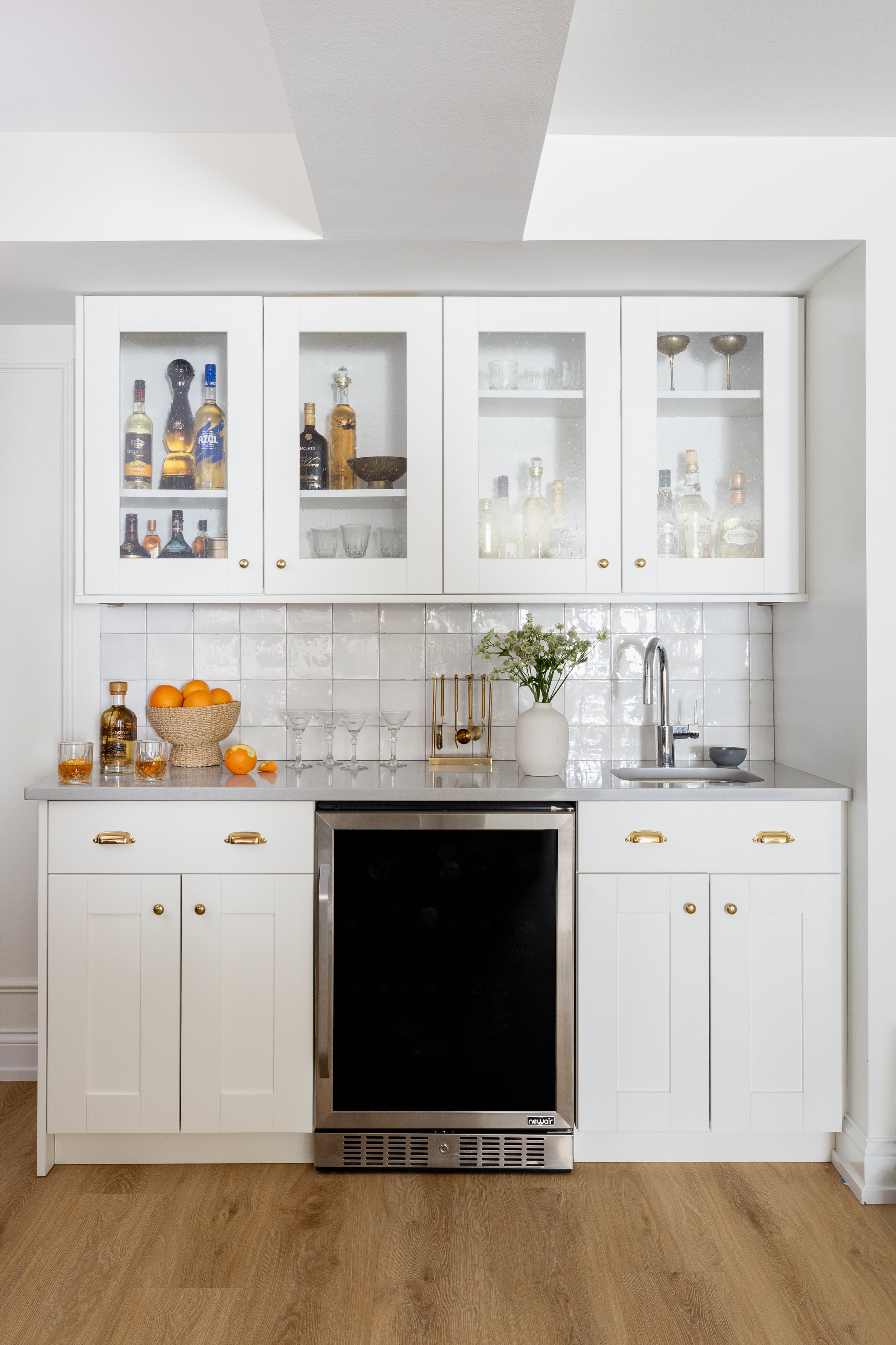 White kitchen cabinet with glass doors, containing liquor bottles and glassware, above a countertop with oranges in a basket, a white vase with flowers, and a small black appliance, with wooden flooring.