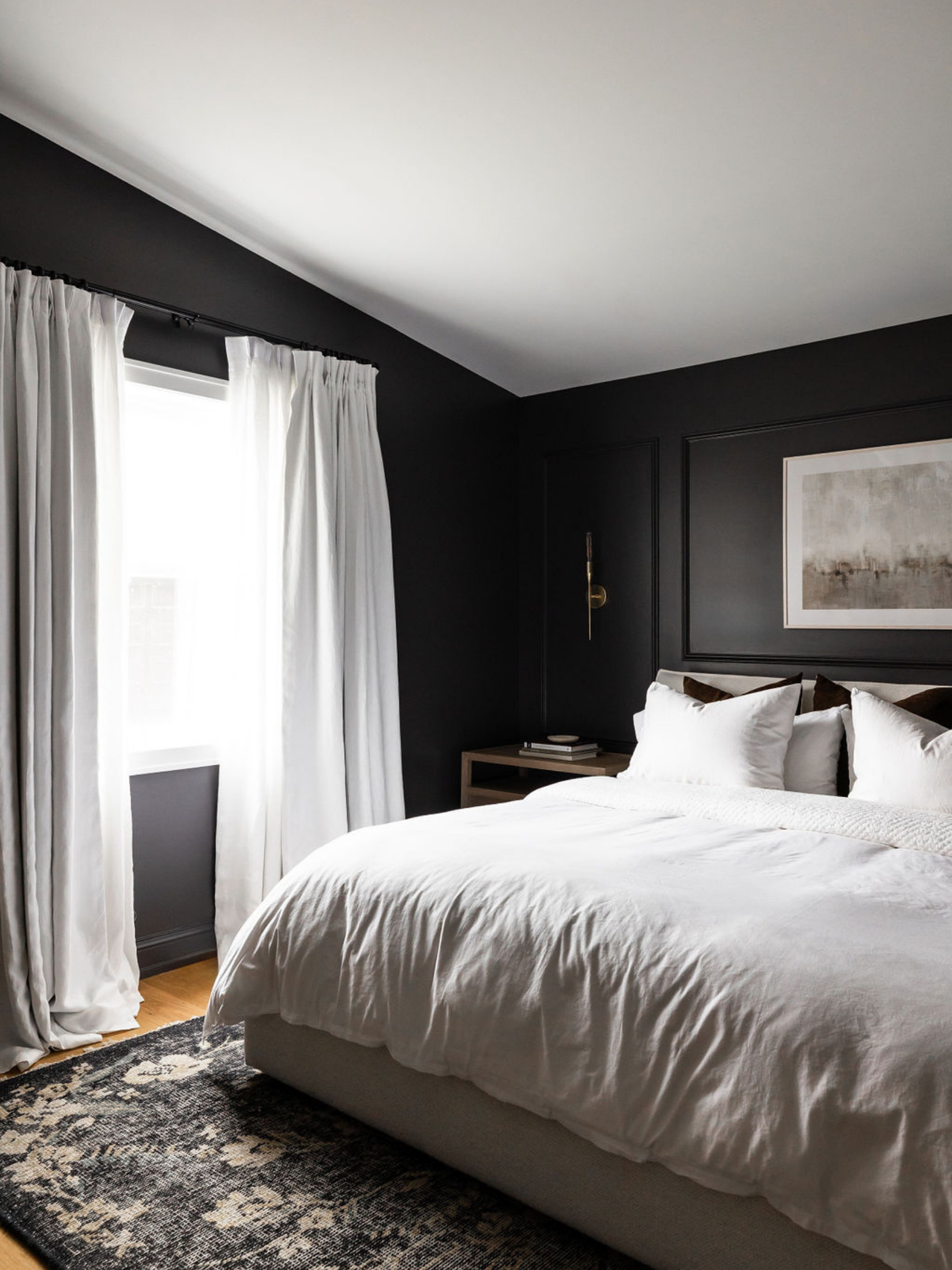 A bedroom with a large bed covered in white bedding, positioned near a window with white curtains. The wall behind the bed is painted dark, and there is a piece of artwork hanging above the bed. There is a bedside table with a lamp and some books.