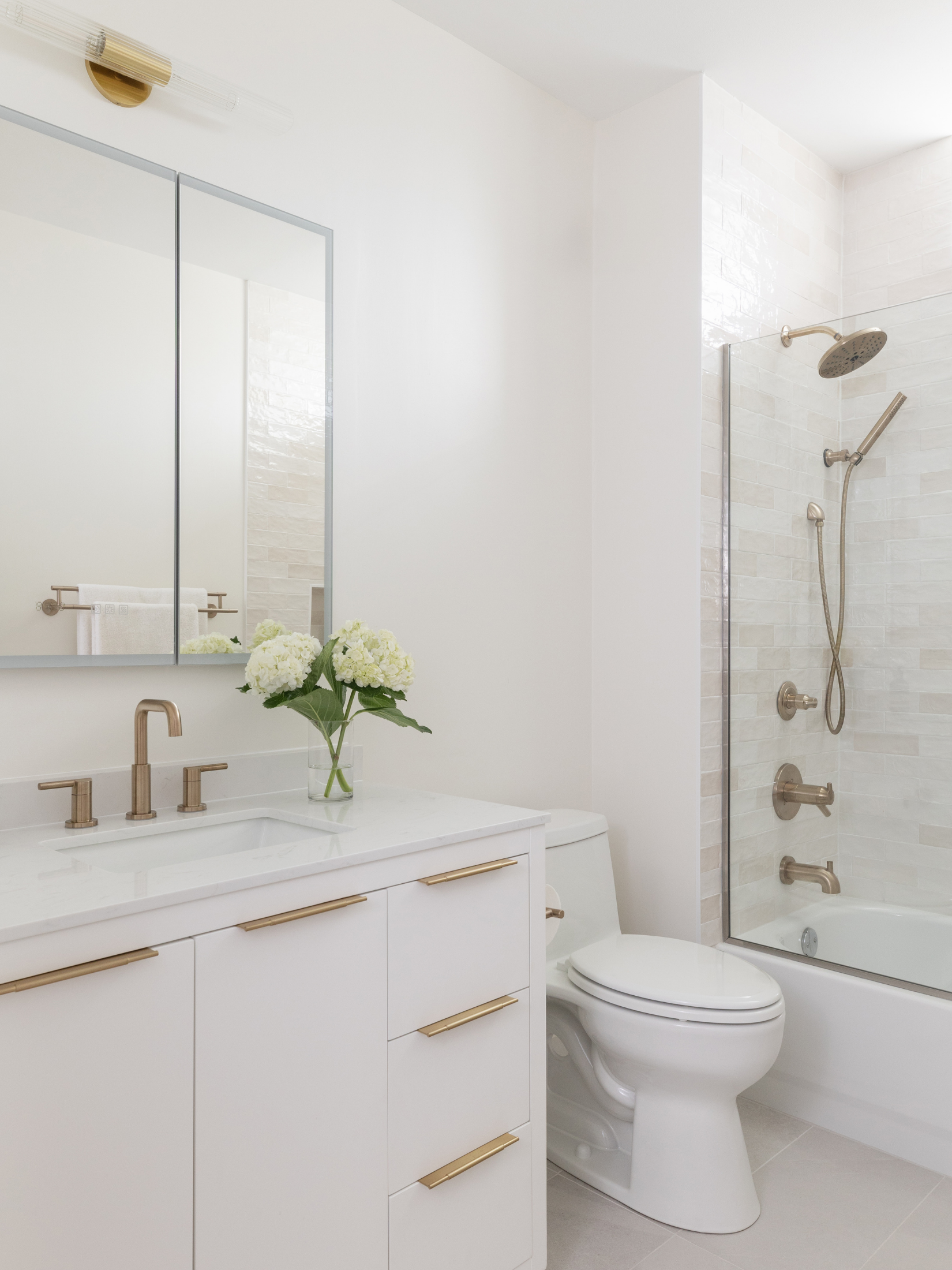 Modern bathroom with a white vanity, gold handles, a flower vase, mirror, toilet, and a shower with beige tiles and a dual showerhead.