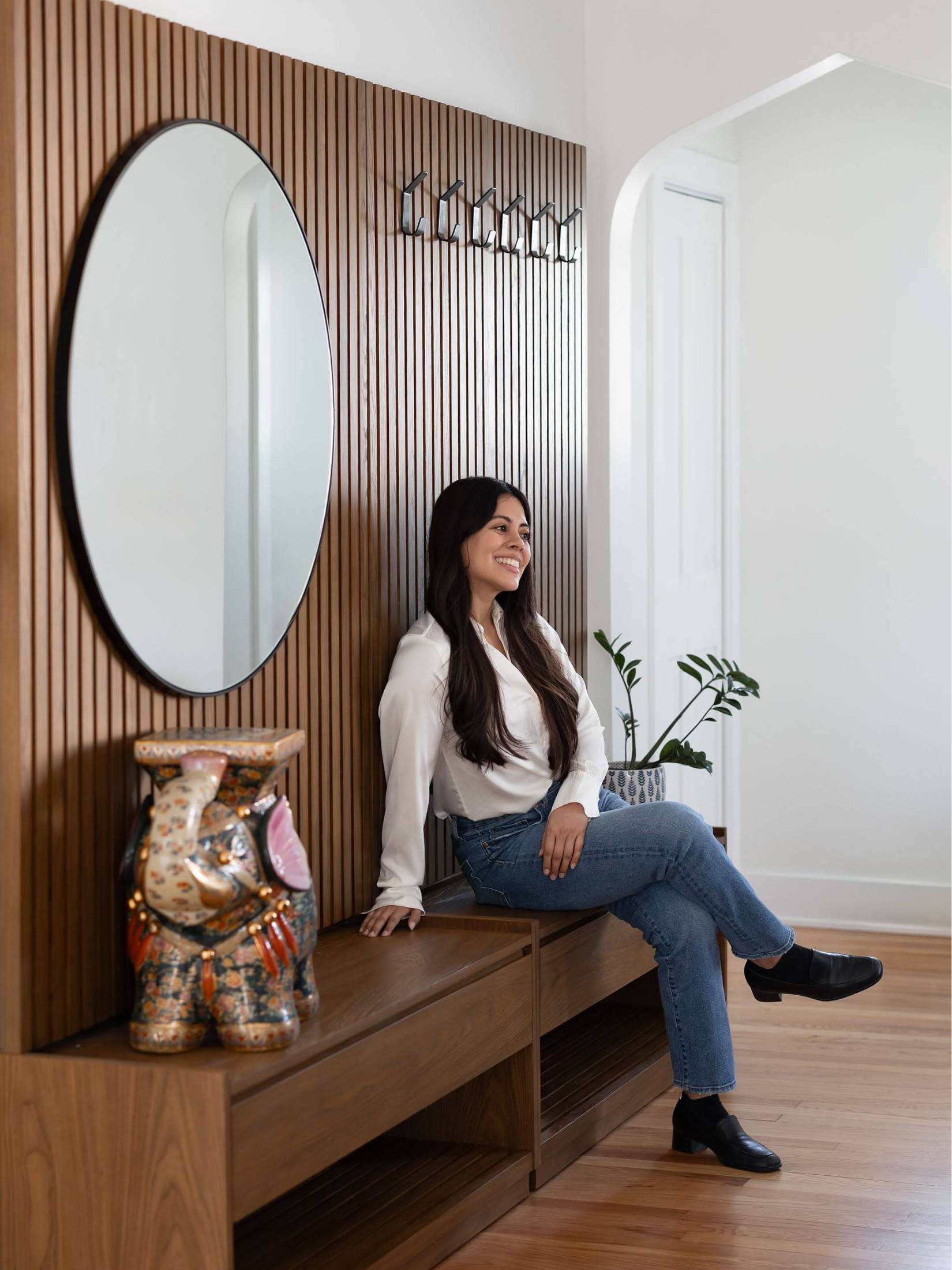 A woman with long dark hair in a white blouse and jeans smiling while sitting on a wooden bench in a modern room with a wooden slat wall, a round mirror, a potted plant, and a decorative pair of colorful, patterned boots.