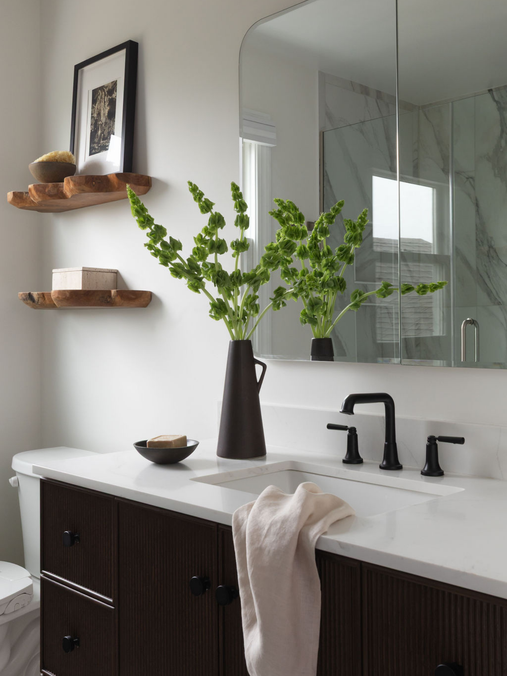Bathroom vanity with dark wood cabinets, white countertop, black faucet, a large mirror with a marble wall behind, and a black vase with green flowers. Two wooden shelves with decor items and a framed picture are on the wall.