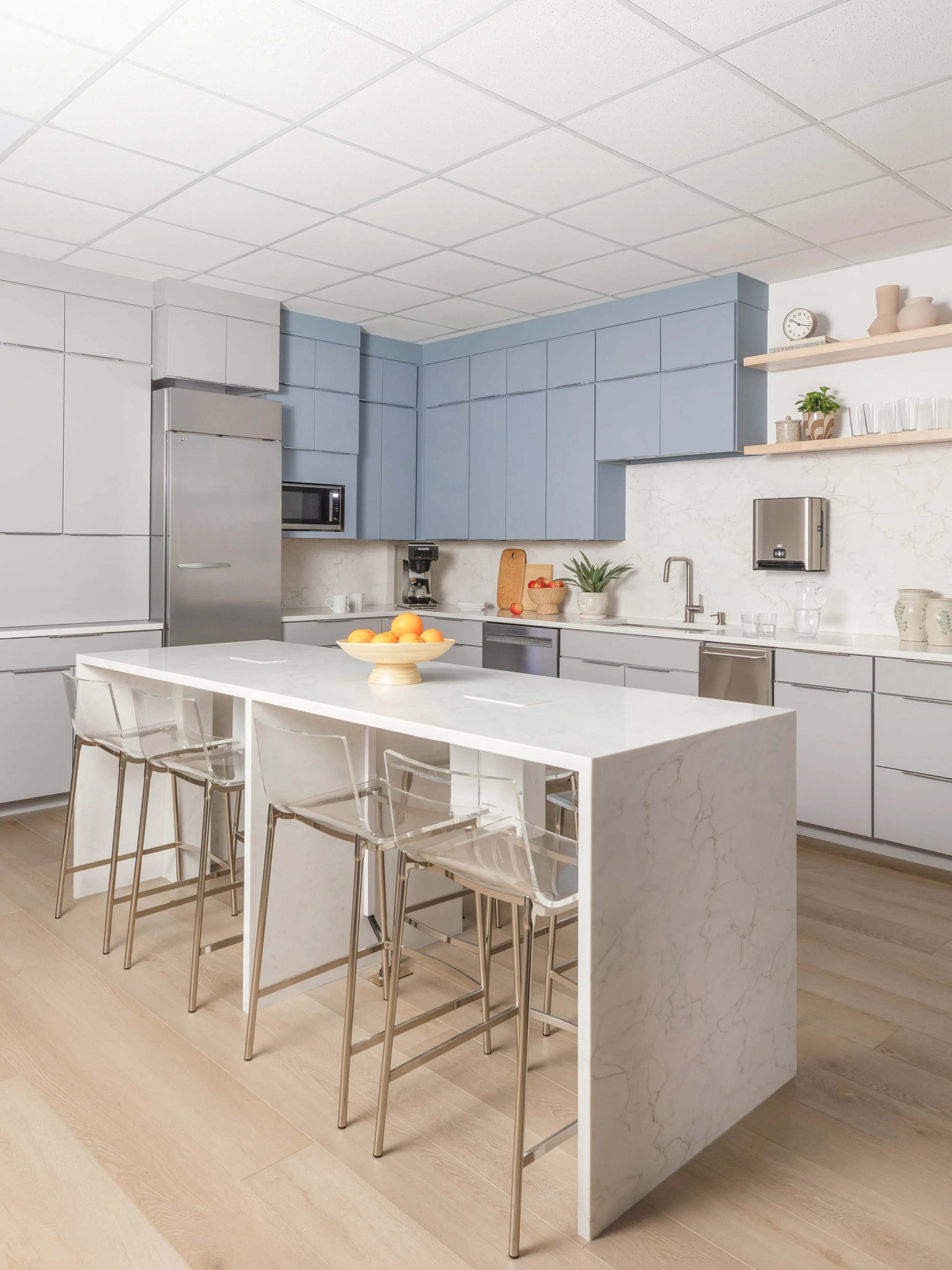Modern kitchen with white and blue cabinetry, marble island, light wood flooring, and open shelving with vases, clock, and glassware.