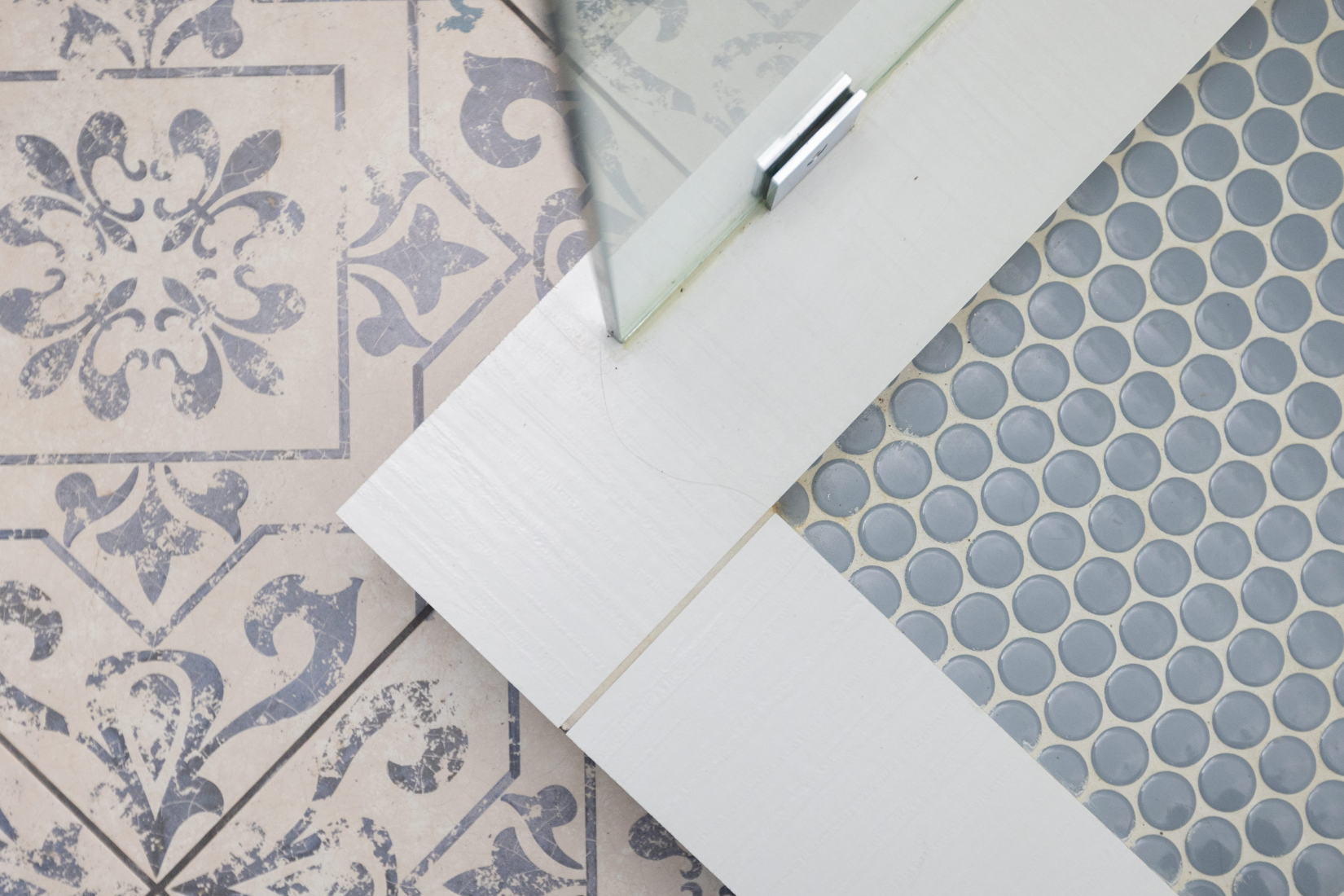 Top view of a tiled floor with contrasting patterns: one section has vintage floral and geometric patterned tiles in beige and gray, while another section has gray circular tiles, separated by a white wooden or faux wood trim.
