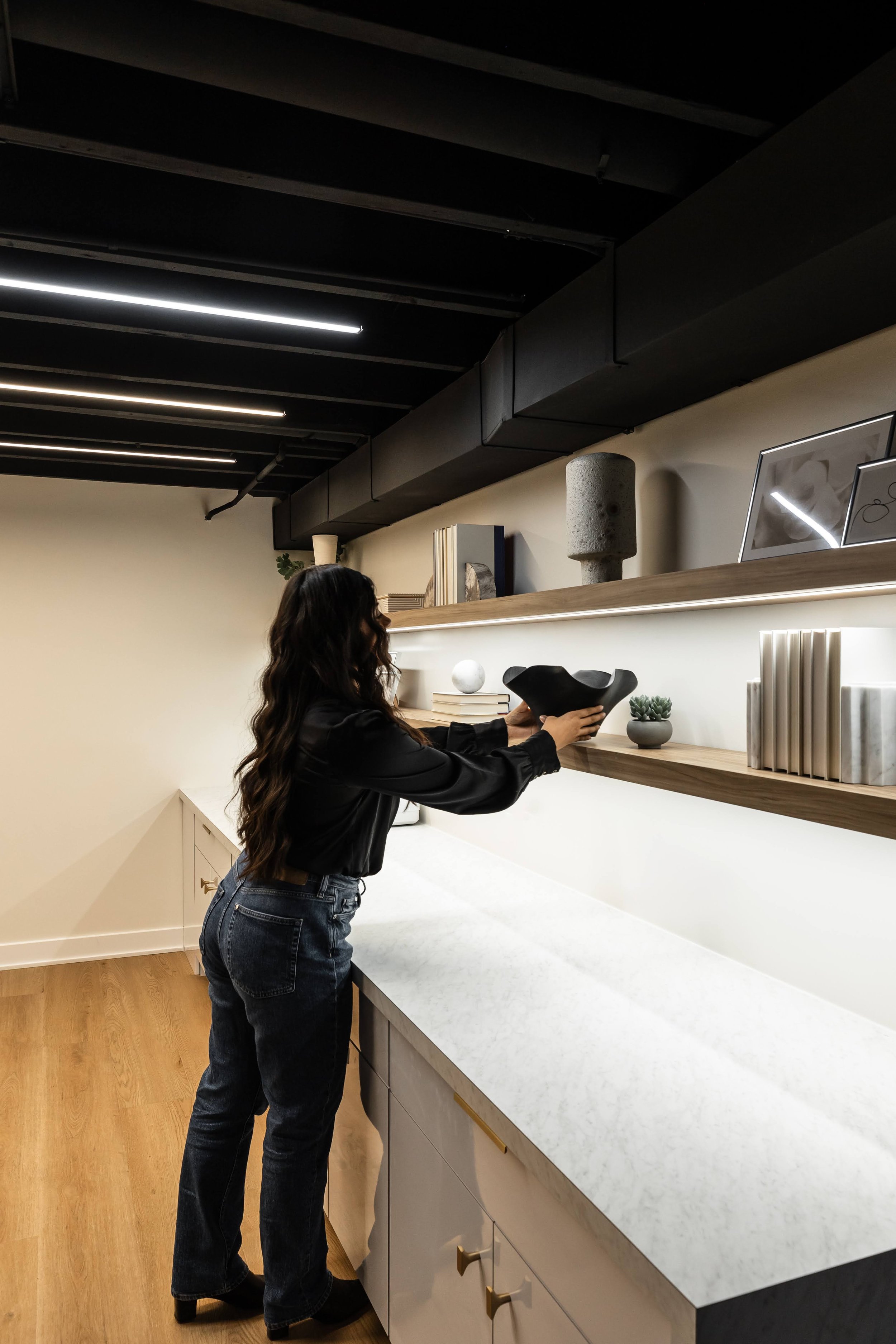 A woman with long dark hair, wearing a black shirt and jeans, is arranging black and white decorative objects on a wooden shelf above a white kitchen countertop. The kitchen features beige cabinets with brass handles and a wood floor.