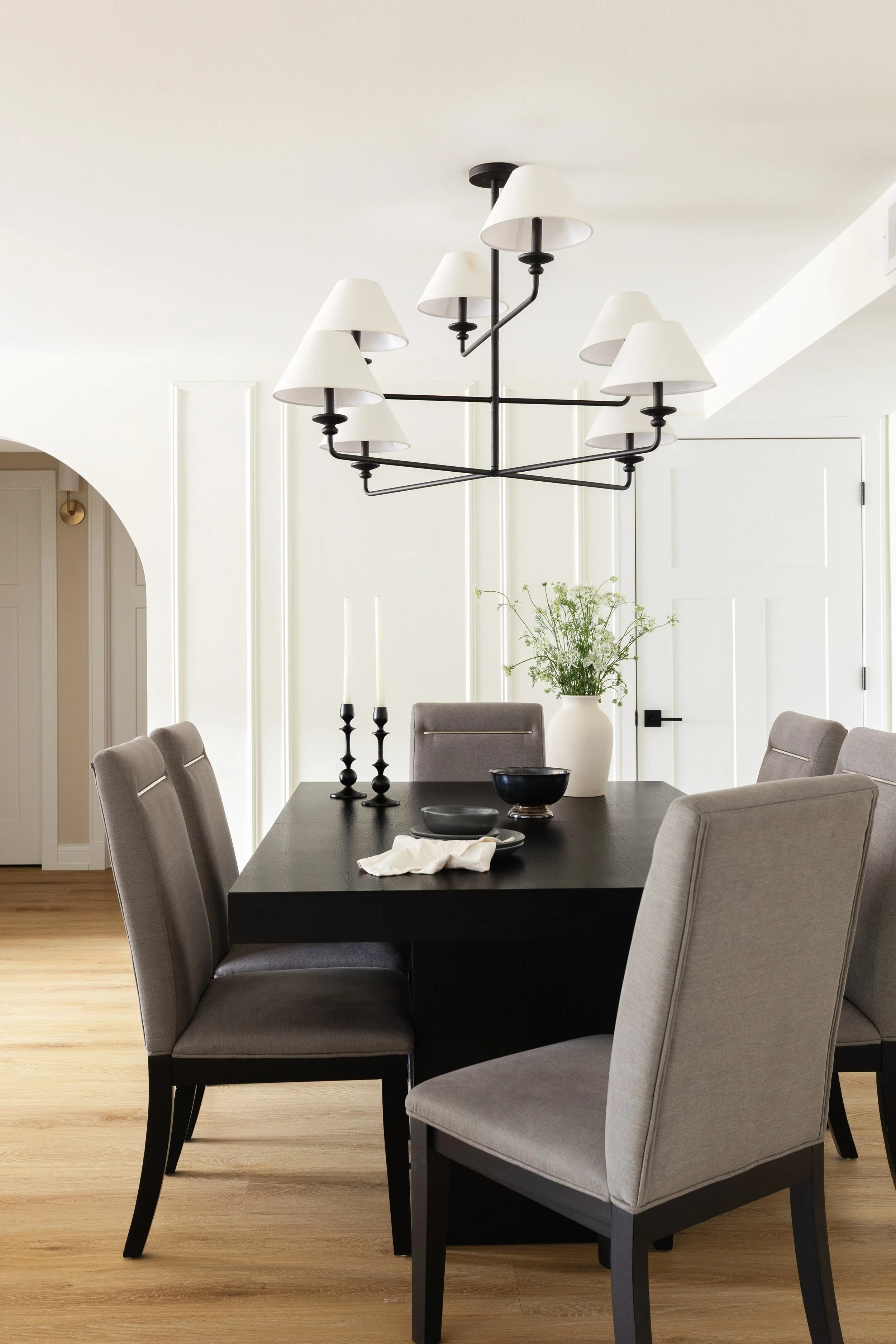 Modern dining room with a black rectangular table surrounded by eight gray upholstered chairs, decorated with black candlesticks, a black bowl, a white napkin, and a large white vase with green plants. A black and white chandelier hangs from the ceil