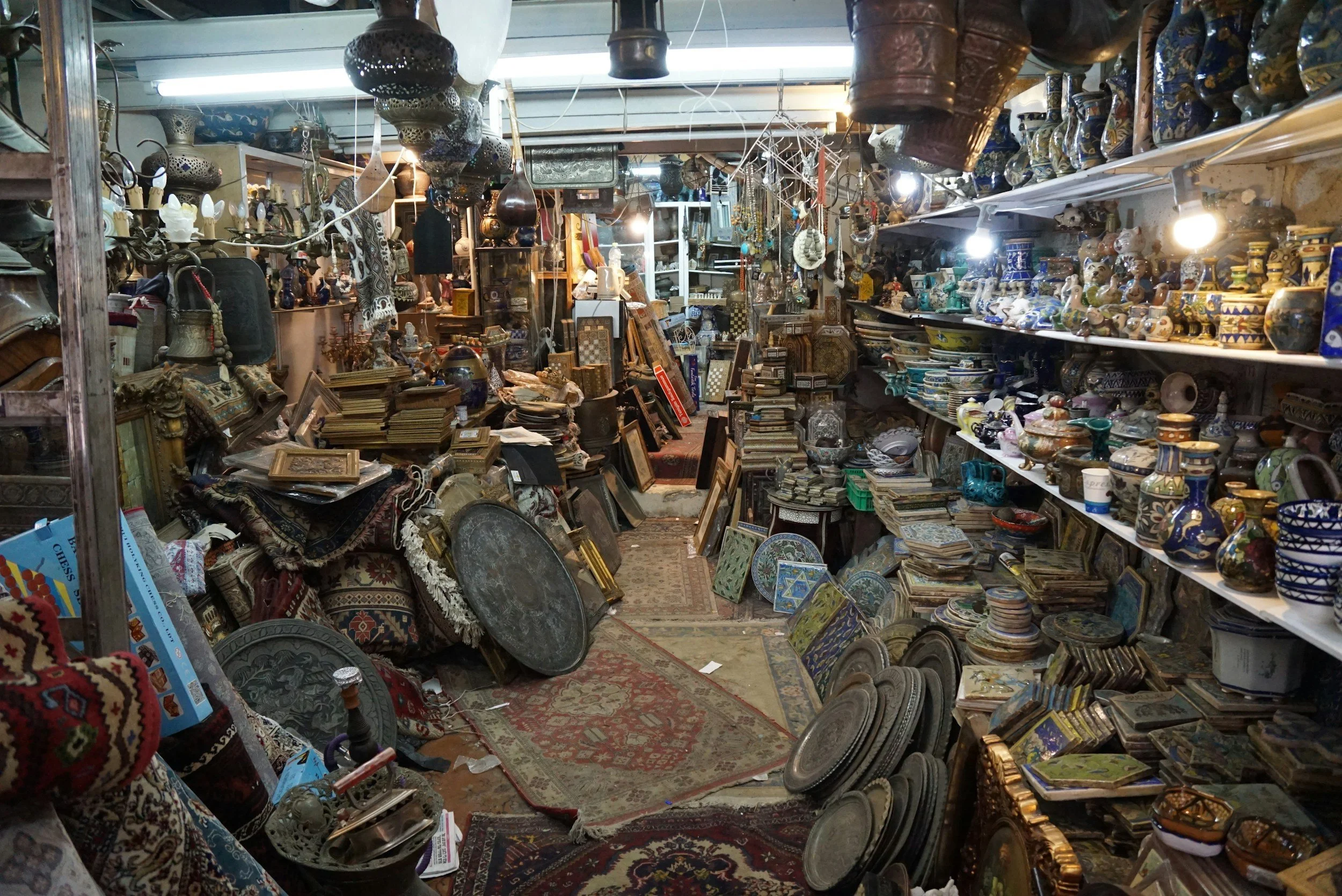 Interior of an antique store filled with shelves and tables stacked with various vintage items, including pottery, plates, artwork, rugs, and decorative objects in a cluttered arrangement.