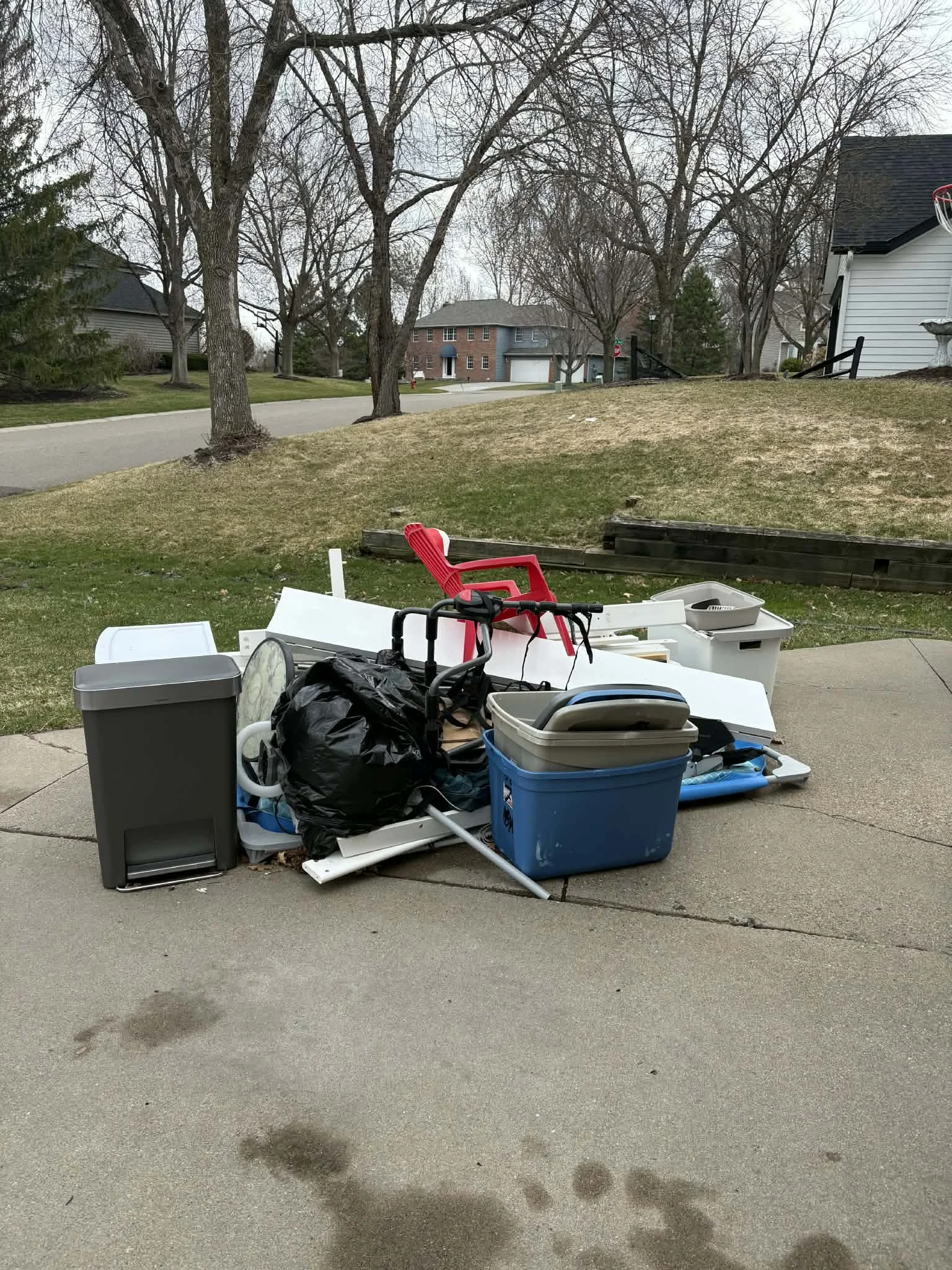 Pile of yard sale items on driveway including plastic bins, a red chair, trash bags, a black bag, and other assorted objects, with trees and houses in the background.