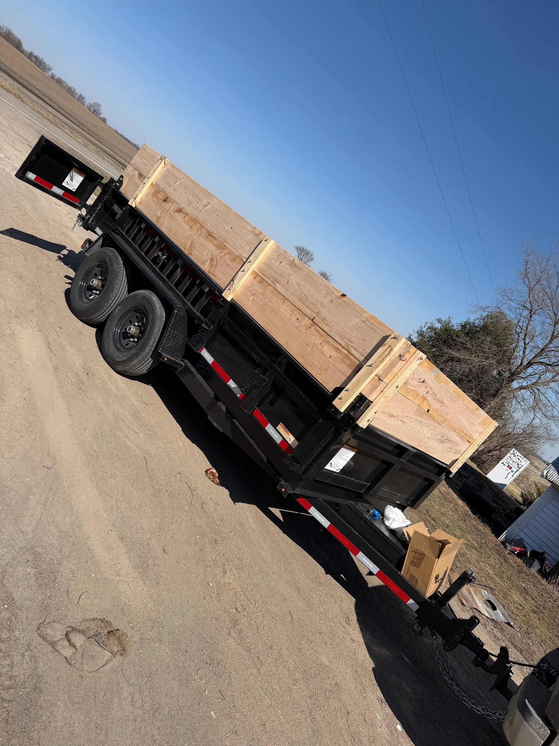 A black flatbed trailer with wooden side rails parked on a dirt lot under a clear blue sky.