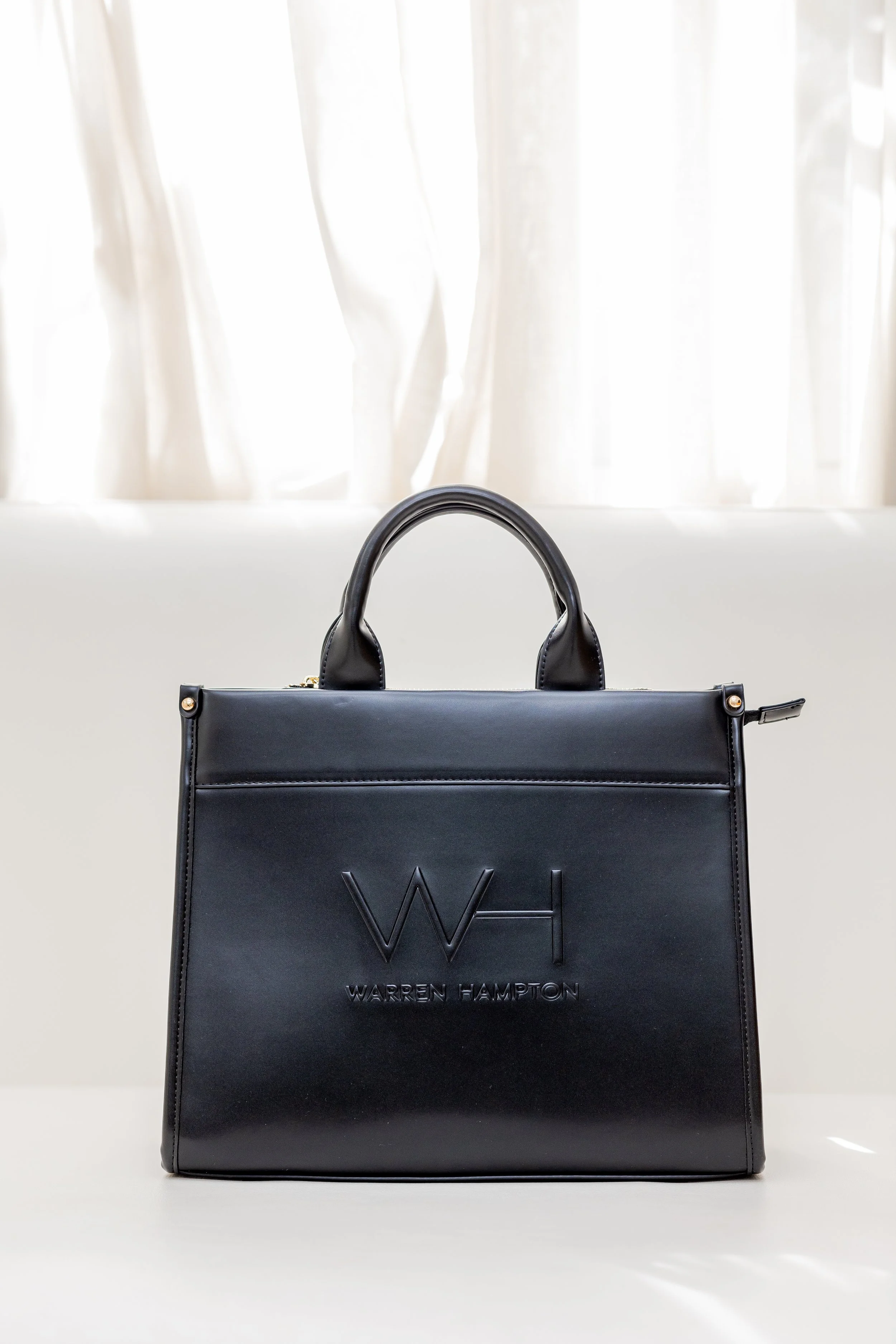 A black handbag with the letter 'W' embossed and the name 'Warren Hampton' on it, placed on a white surface with white curtains in the background.