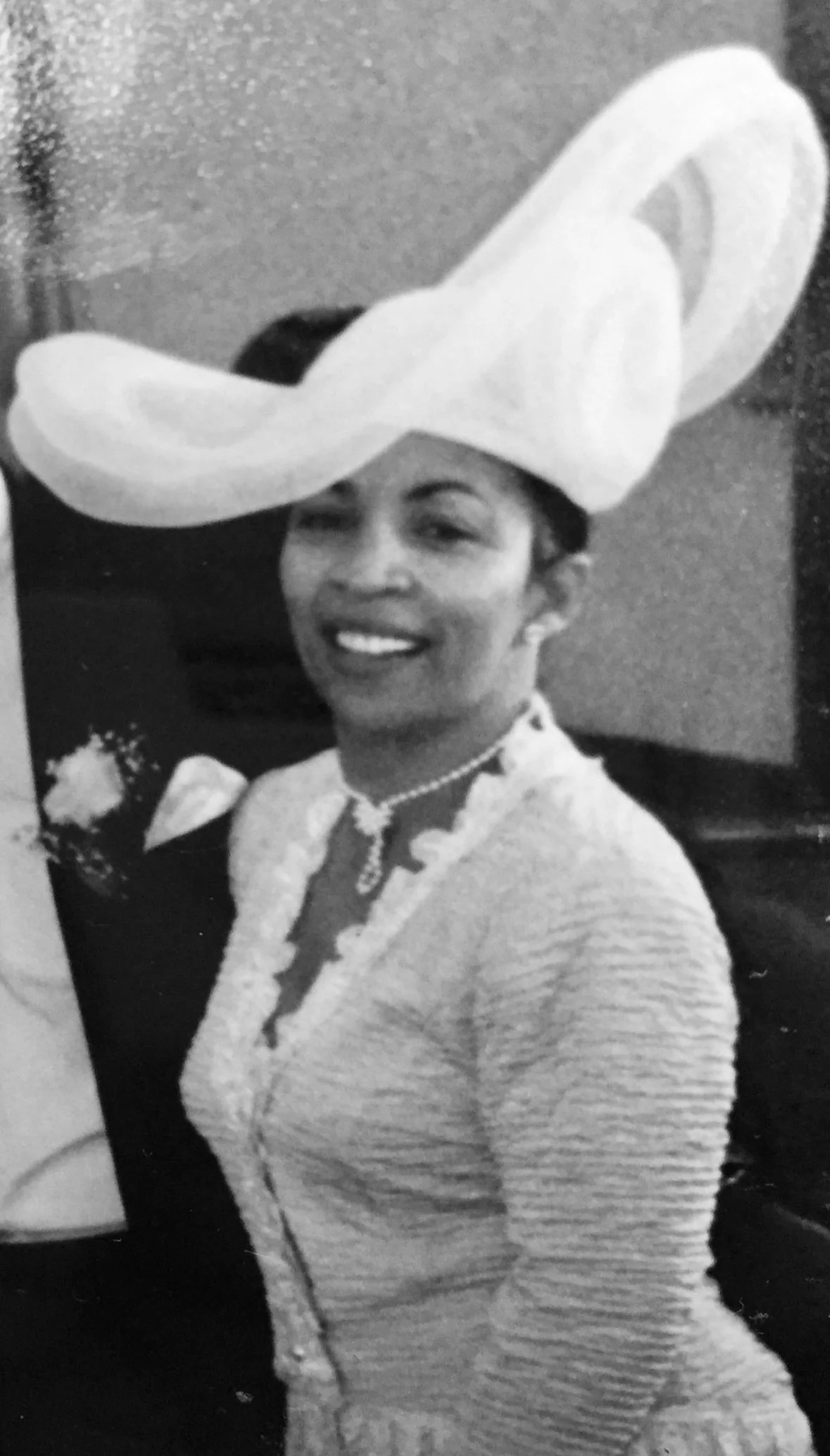 Black and white photo of a woman smiling, wearing a large hat, a pearl necklace, and a cardigan, possibly at a social event.