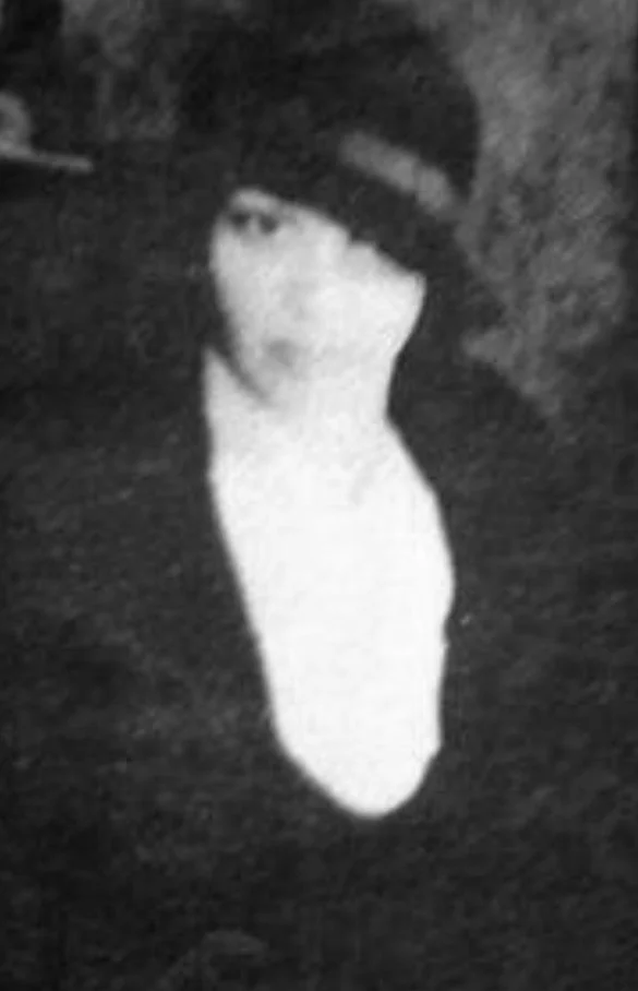 A person with dark hair wearing a dark cap and white shirt, looking at the camera in a low-light setting.