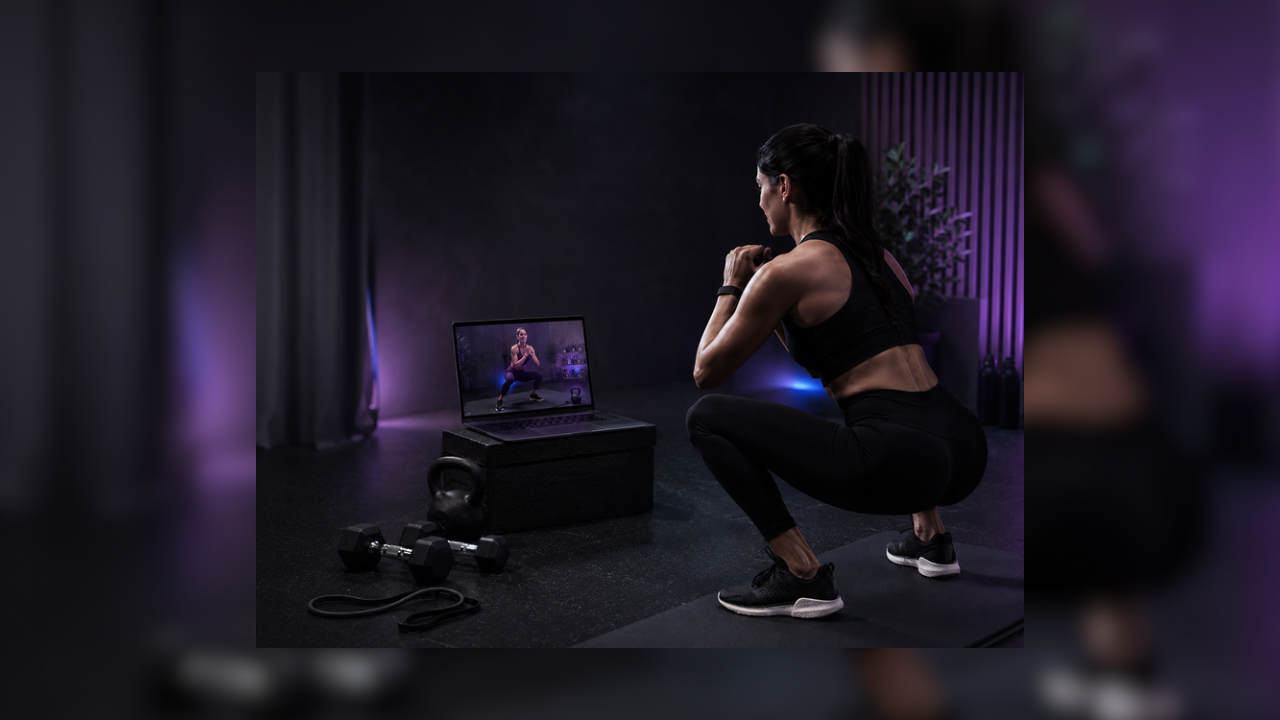 A woman is doing squats in a gym with a laptop showing an instructor, with dumbbells and a punching bag nearby.
