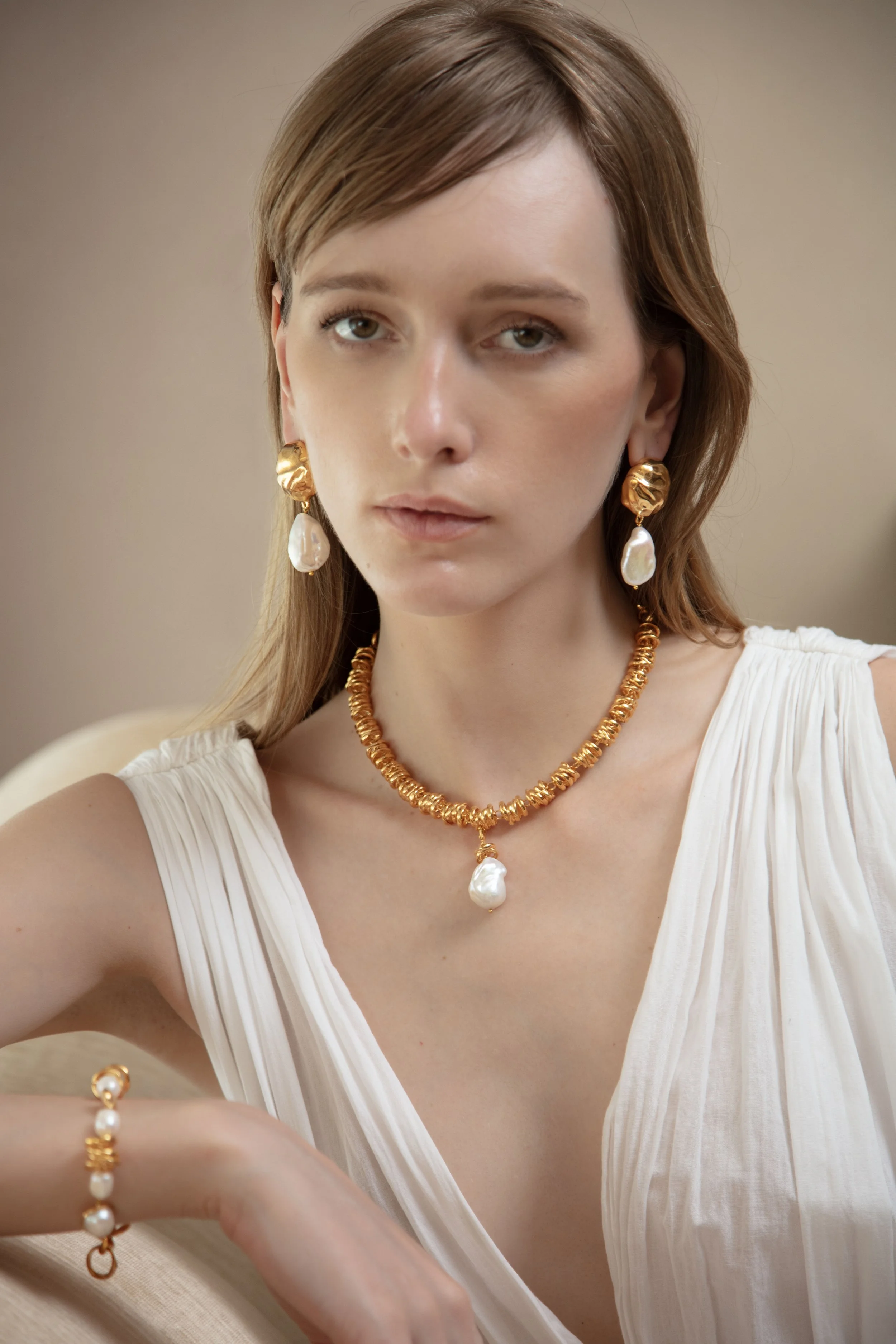 A woman with light brown hair wearing gold and pearl jewelry, including earrings, a necklace, and a bracelet, dressed in a white pleated sleeveless top, sitting against a neutral background.