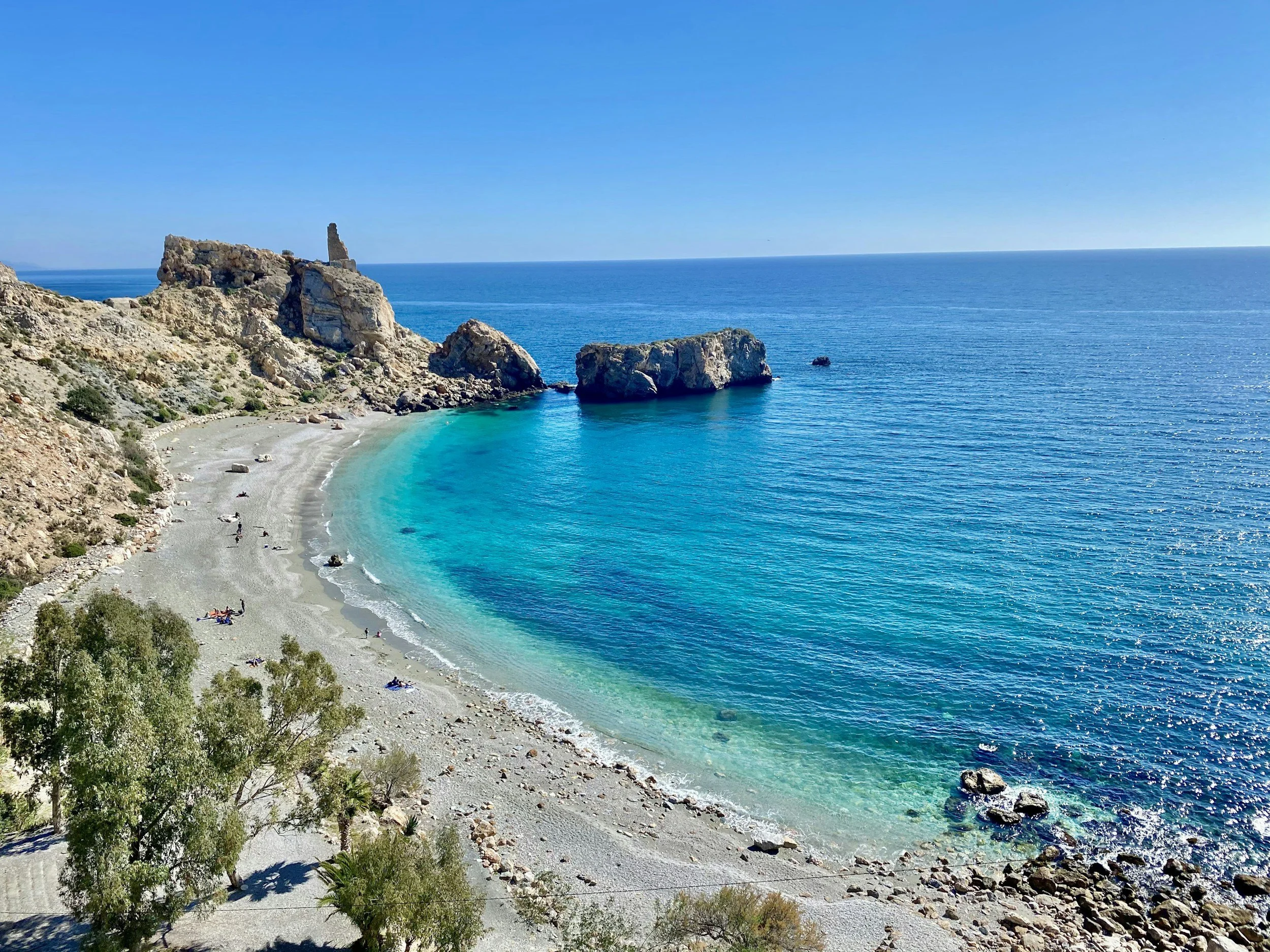 A scenic view of a beach with clear blue water, surrounded by rocky cliffs and sparse vegetation, with a few people relaxing on the sandy shore.