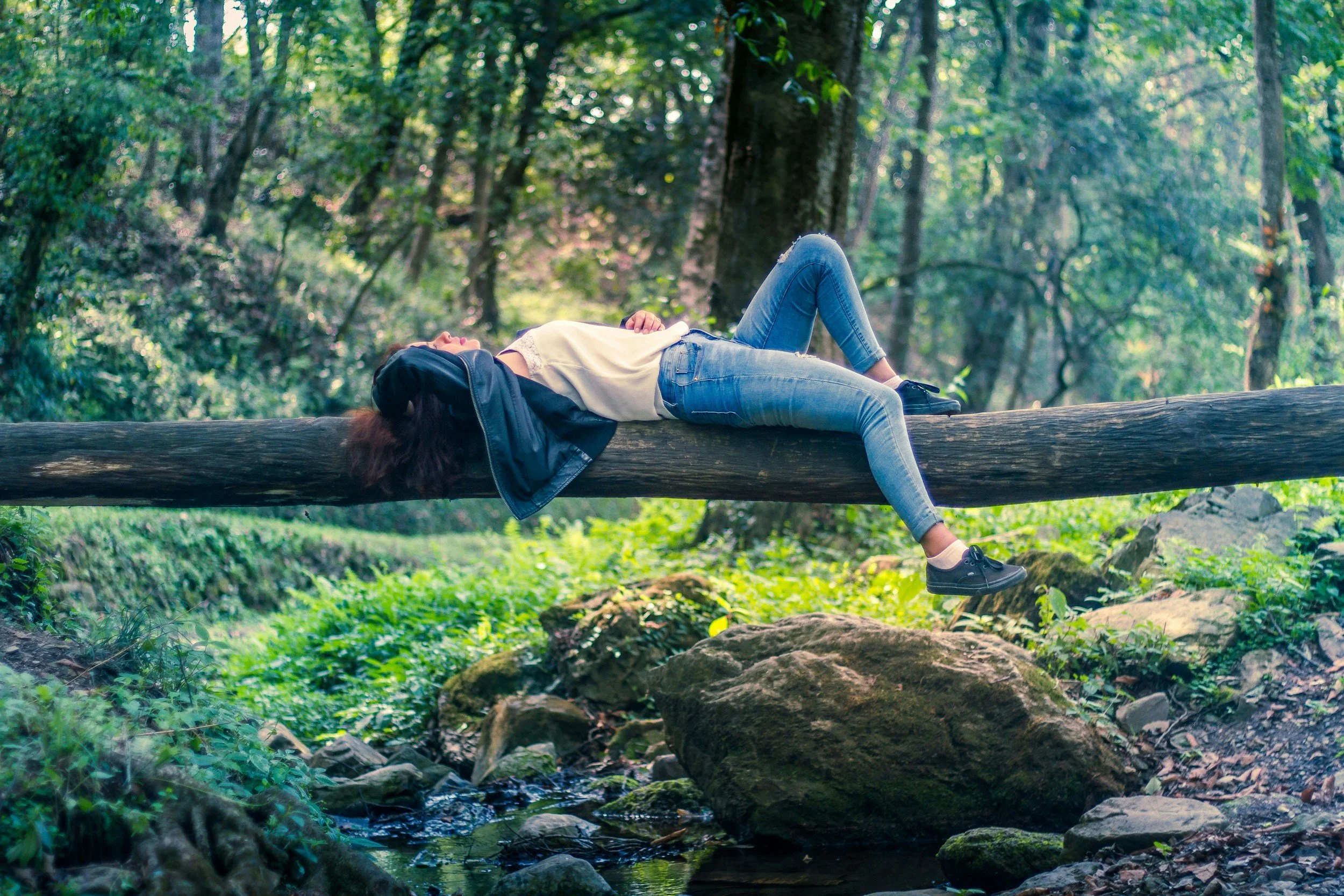 A woman lying on her back on a fallen tree log in a forest, with her head and hair resting on the log and her legs hanging off the side, surrounded by green trees and rocks.