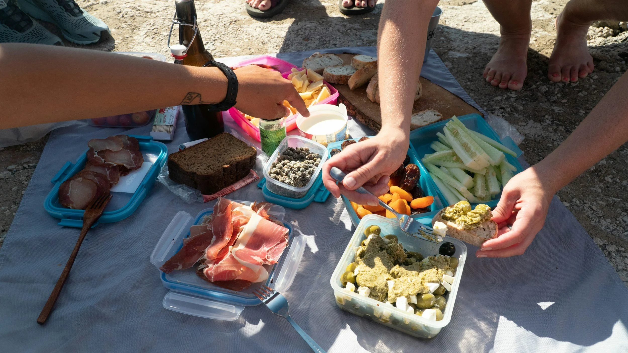 Picnic spread on a gray tablecloth with various foods including sliced meats, bread, cheese, vegetables, olives, and spreads, with people serving and preparing food outdoors.