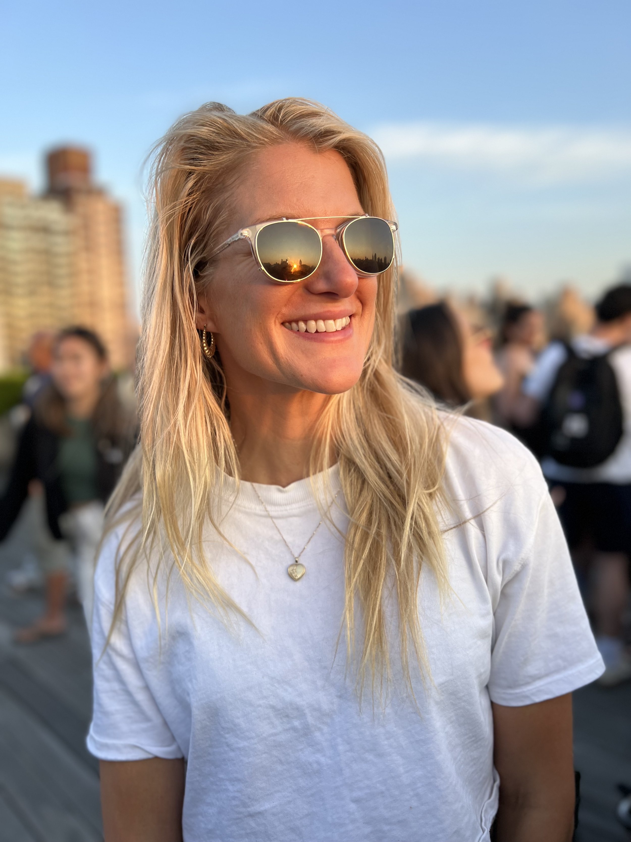 A woman with blonde hair wearing sunglasses, a white t-shirt, and a heart-shaped pendant necklace, smiling outdoors with a cityscape and other people in the background during sunset.