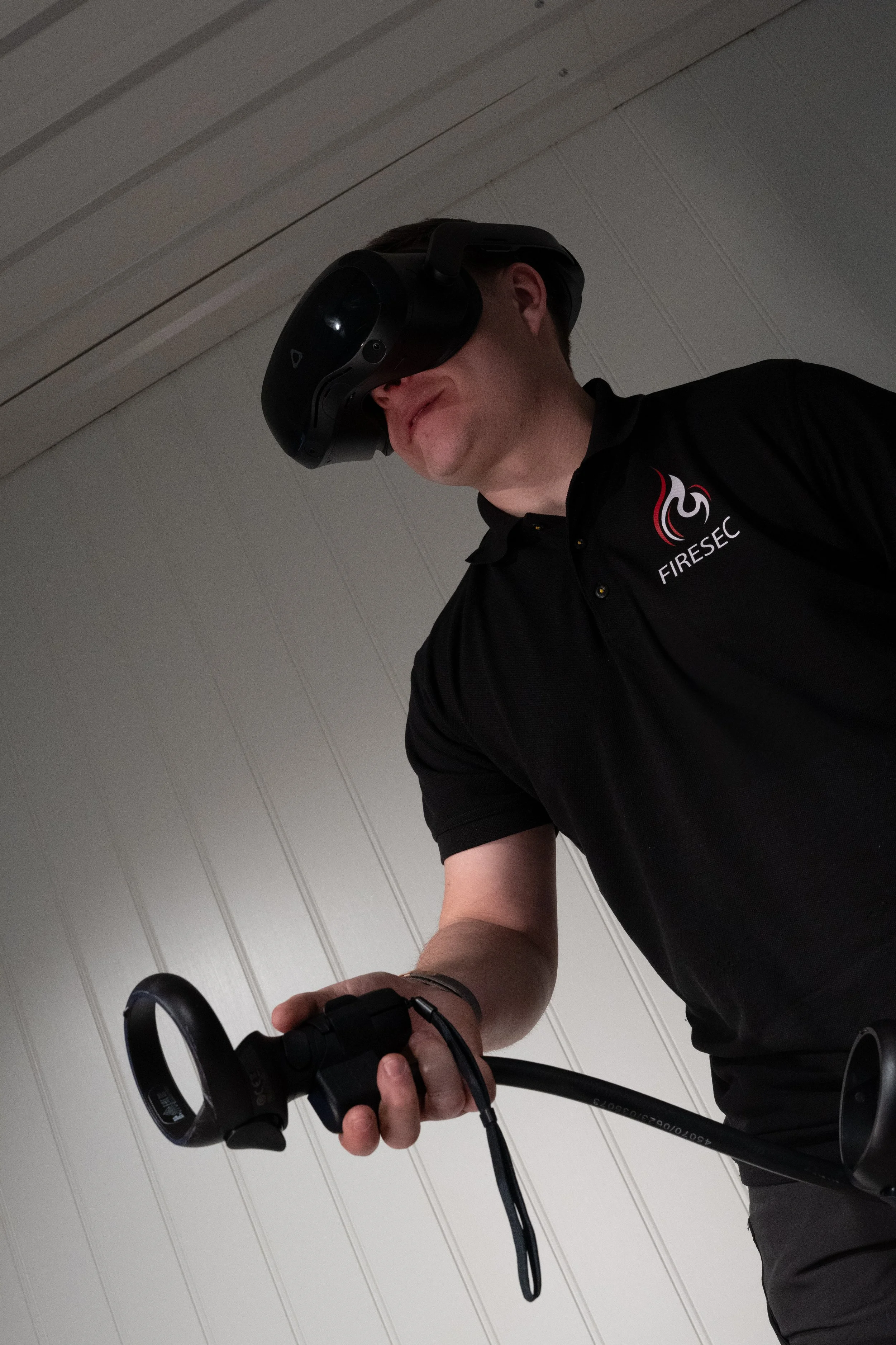 En mann bruker en virtual reality-headset og holder i en styrestang, iført svart skjorte med logoen 'FIRESC' på.