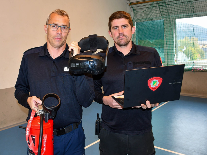 To menn i uniform står i en gymsal med ulike teknologiske verktøy, inkludert en bærbar datamaskin, en drone, og en VR-brille.
