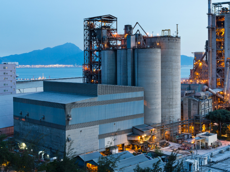 En stor industribedrift med store silos og metallkonstruksjoner ved vannet på kvelden, med fjell i bakgrunnen.