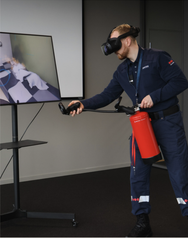 Person i brannmannsuniform bruker VR-hjelm og dynamittøyne til å simulere brannslukking, med brannslukker hengende fra hoften, foran stor skjerm.