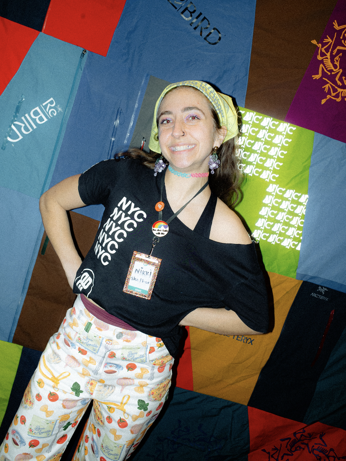 A woman with long brown hair, wearing a yellow bandana, a black T-shirt with 'NYC' repeated multiple times, colorful pants with patterns of bread, tomatoes, and herbs, and rainbow accessories, standing in front of a colorful quilted backdrop with various designs and text.