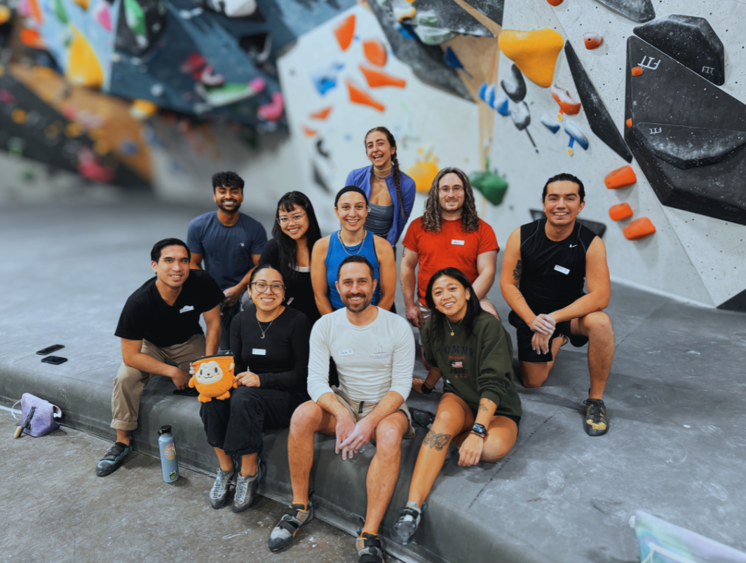 Group of ten people smiling and posing at an indoor bouldering gym, with colorful climbing holds and routes on the wall in the background.