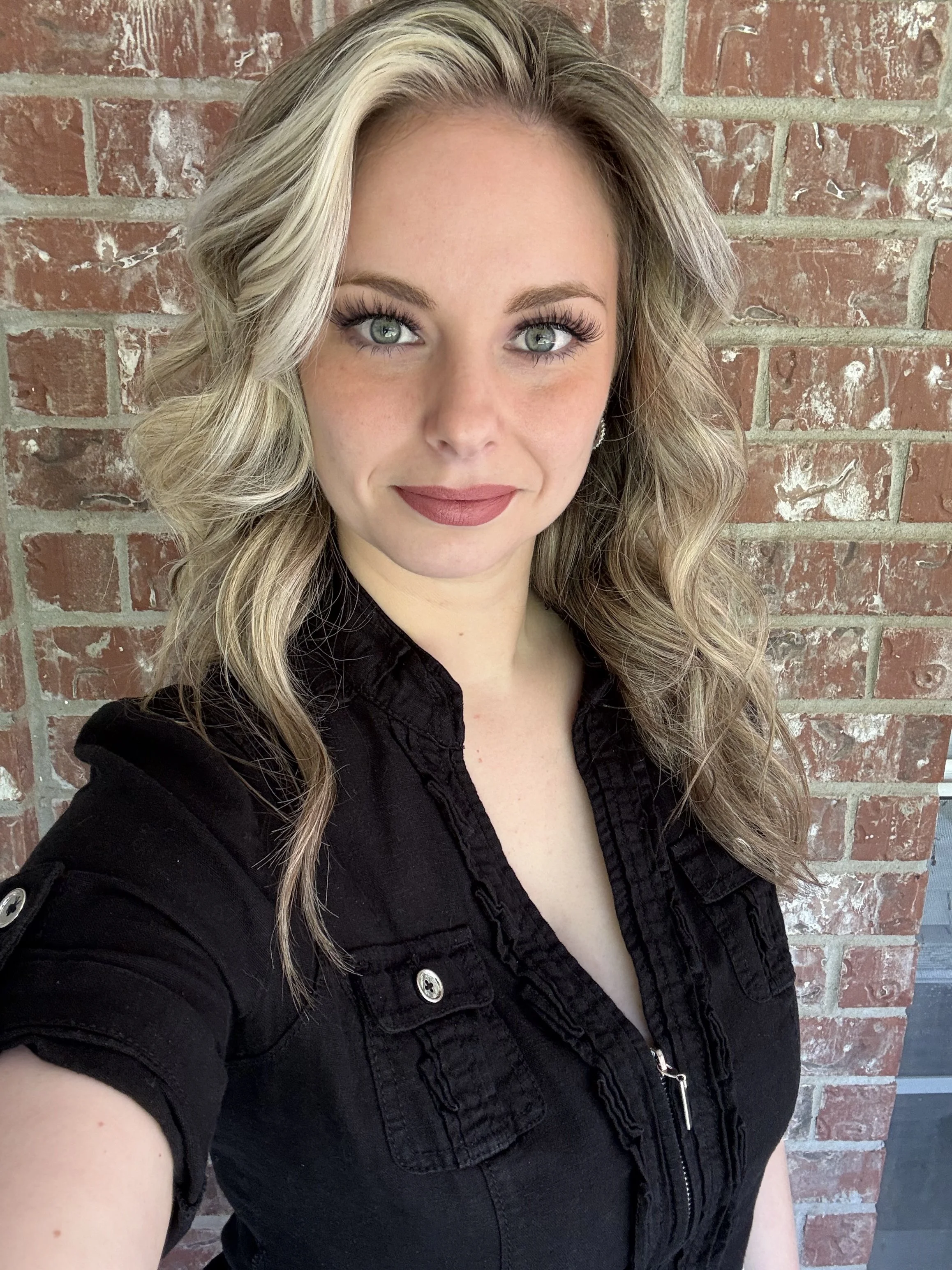 A woman with blonde, wavy hair and blue eyes taking a selfie against a brick wall.