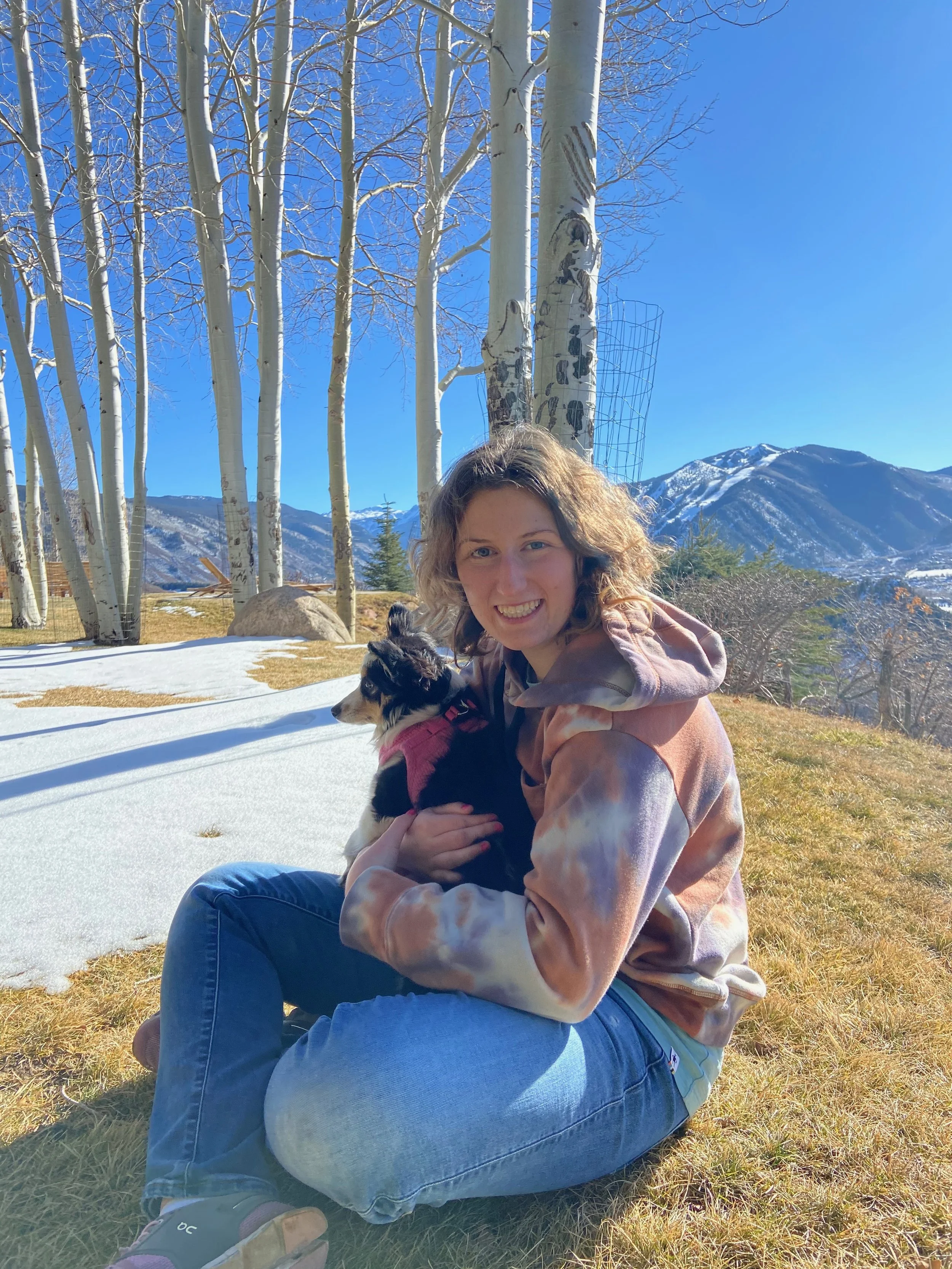 A woman sitting on the grass outdoors, holding a dog, with tall leafless trees, mountain scenery, and blue sky in the background.