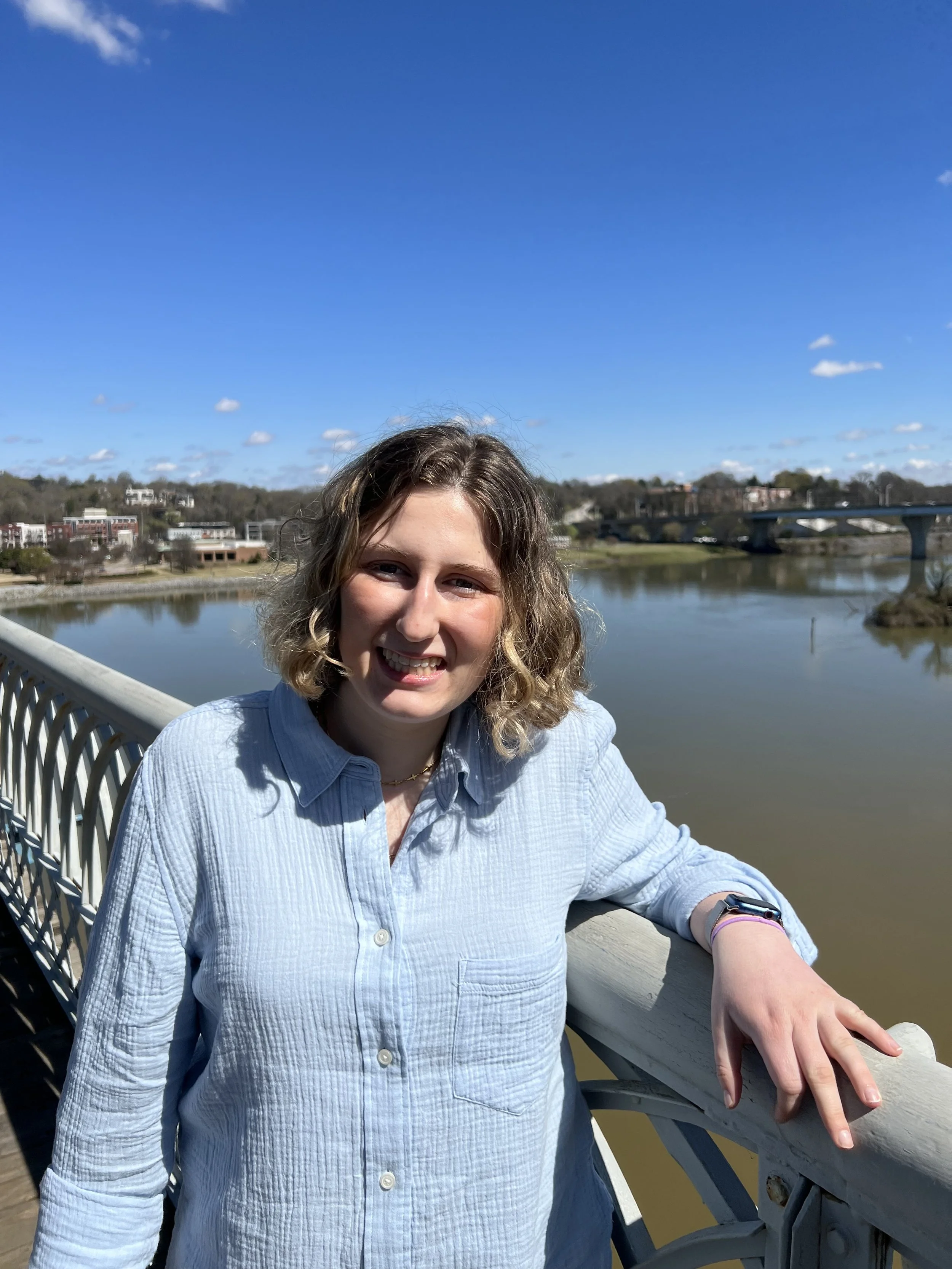 A woman with curly blonde hair smiling as she stands on a bridge beside a river, with a cityscape and a blue sky with scattered clouds in the background.