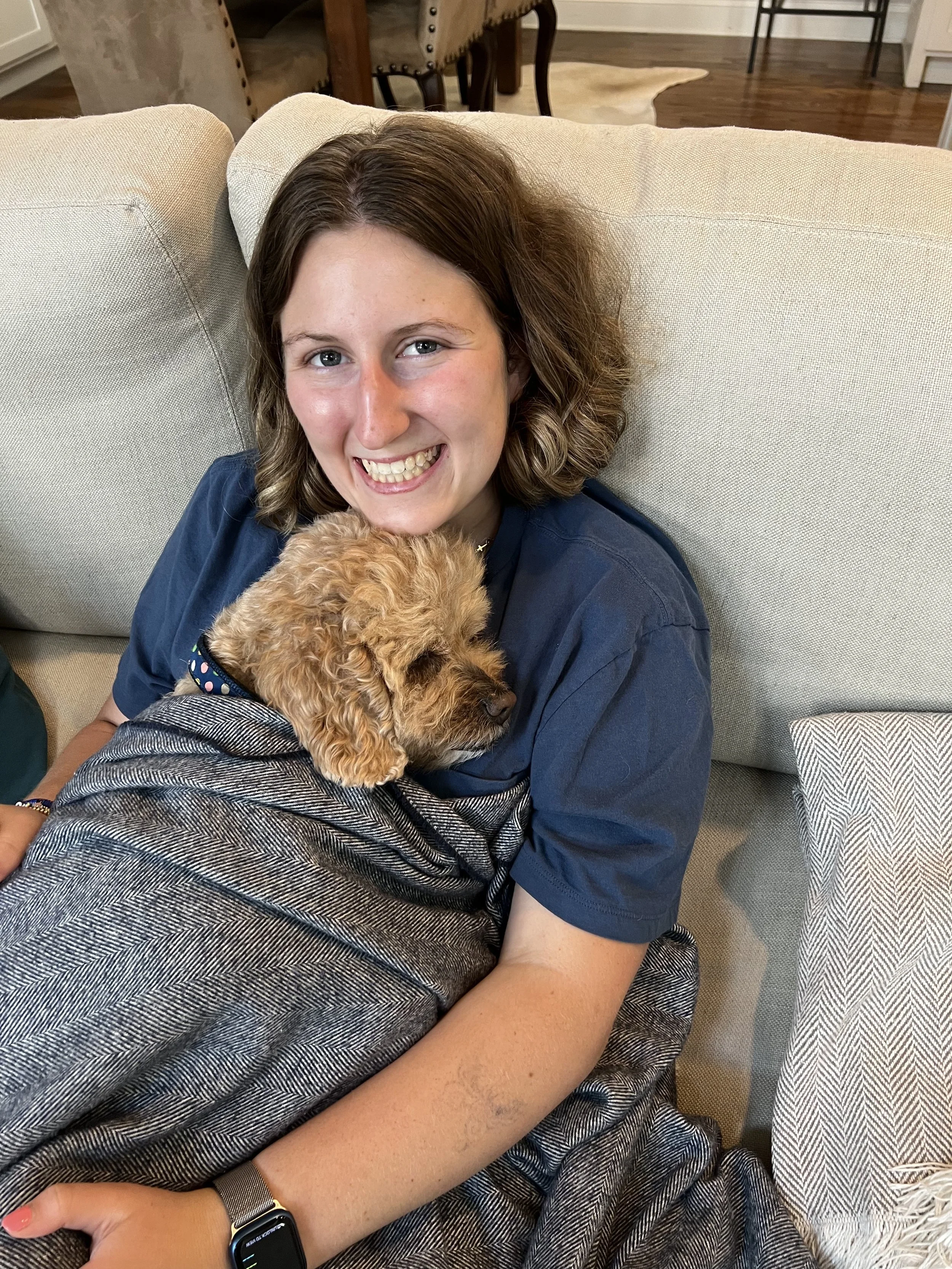 A woman smiling while holding a dog on her lap, sitting on a beige couch in a living room.