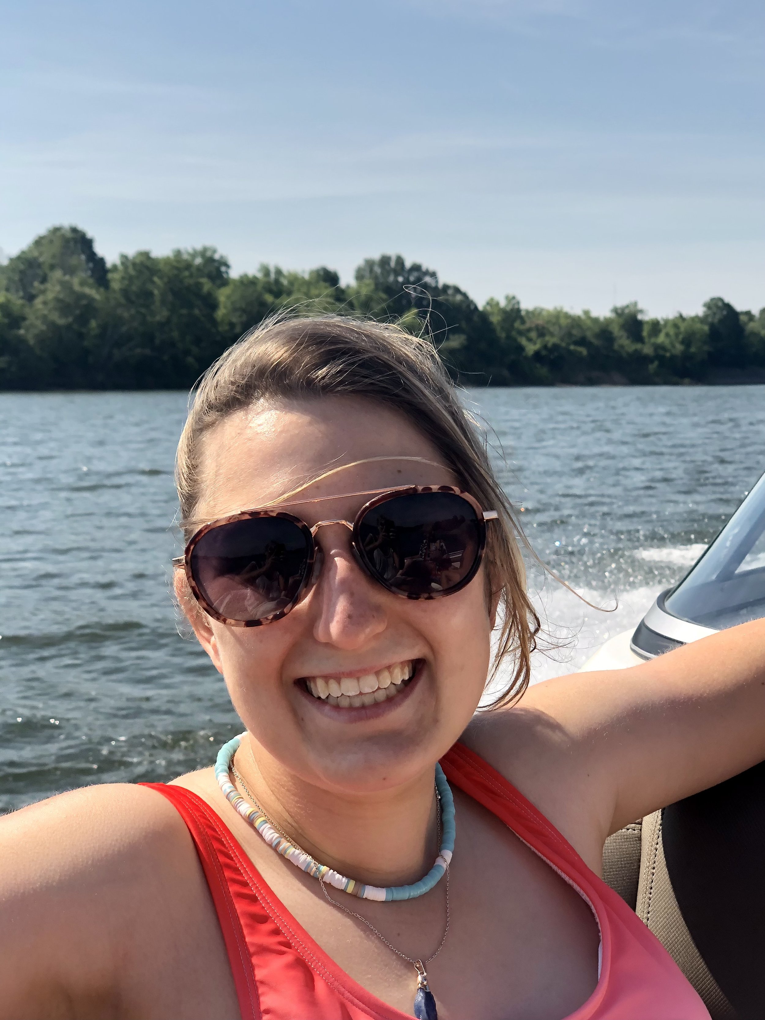A woman smiling while on a boat on a lake with trees in the background, wearing sunglasses, a red swimsuit, and jewelry.