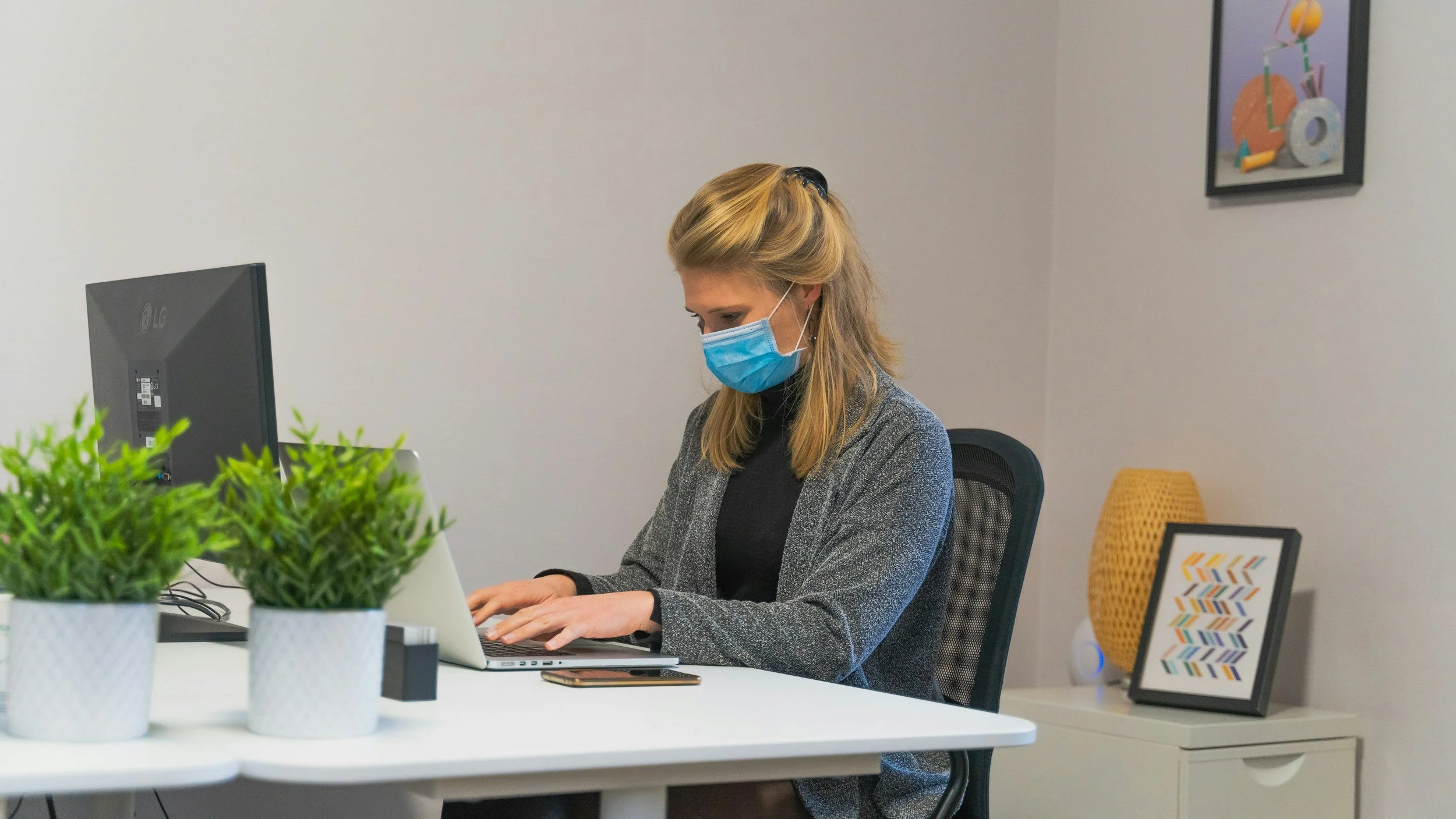 A woman wearing a blue face mask working on a laptop at a white desk with green plants, a computer monitor, framed artwork, and a yellow chair in the background.