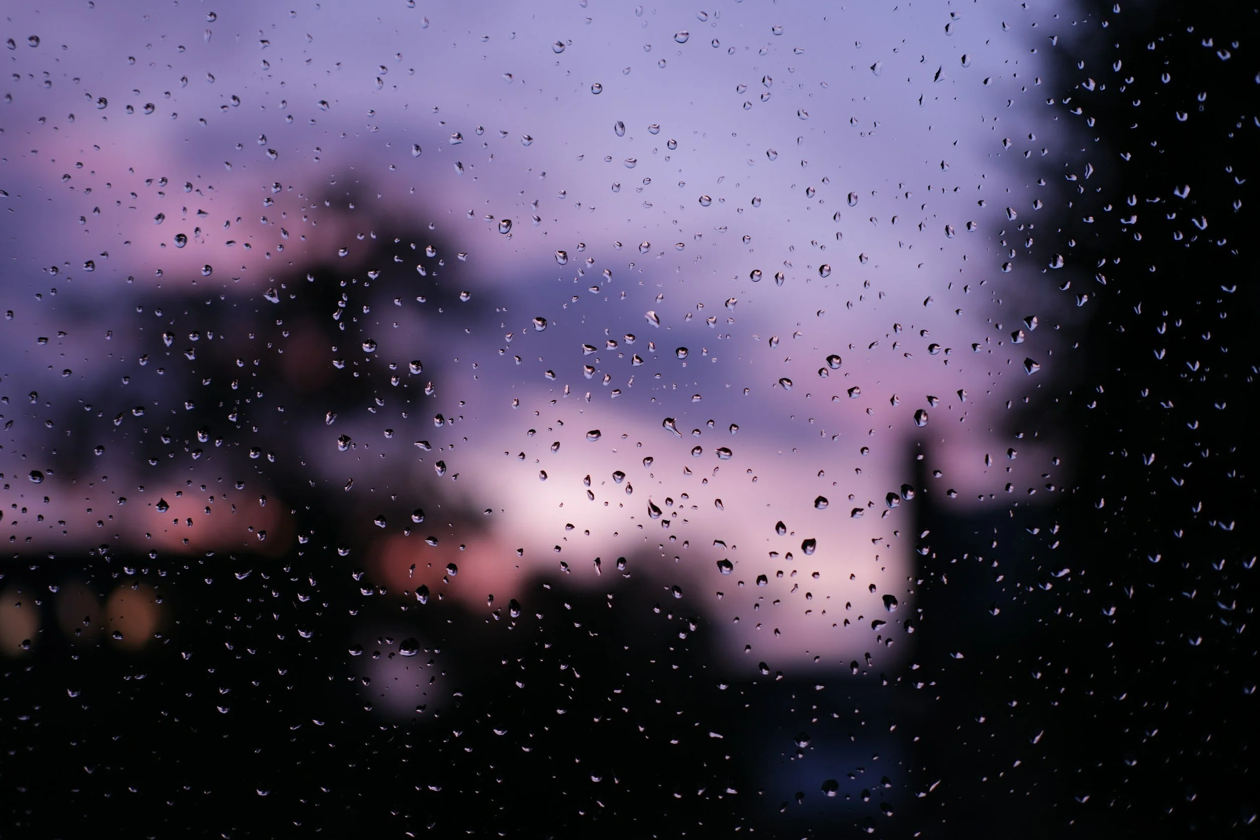 Close-up of a window with raindrops on the glass, blurred trees and purple sky in the background during sunset.